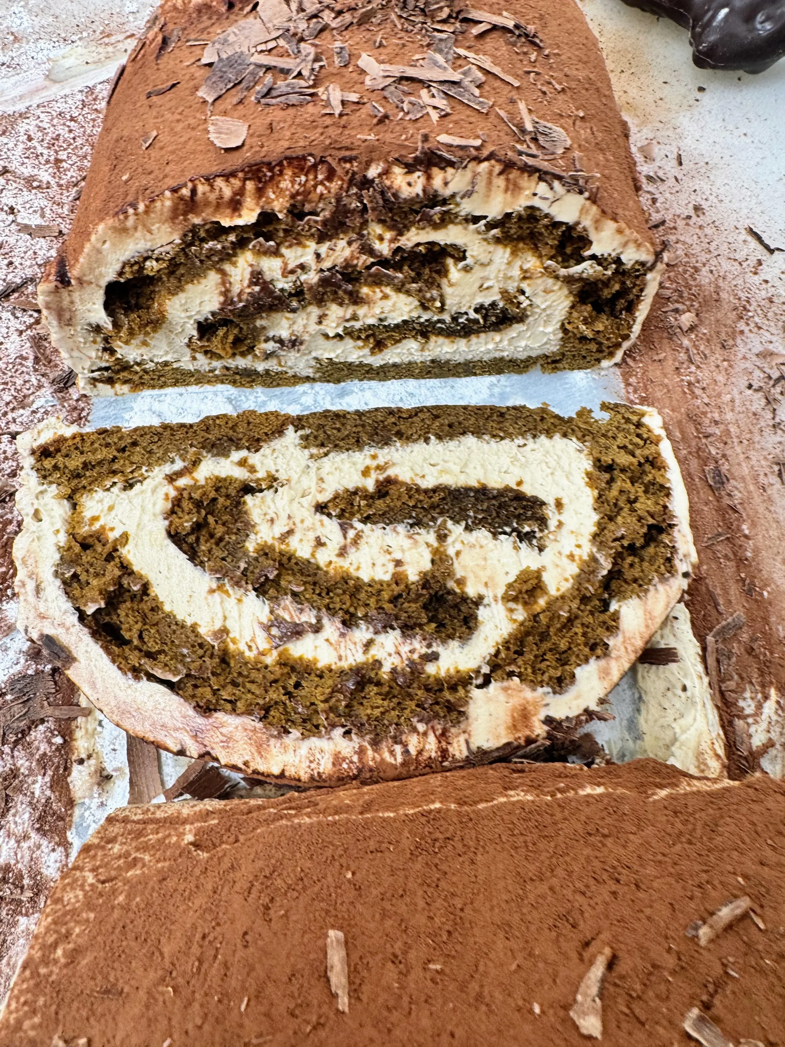 A sliced chocolate and vanilla roll cake with a spiral pattern, topped with cocoa powder and chocolate shavings.