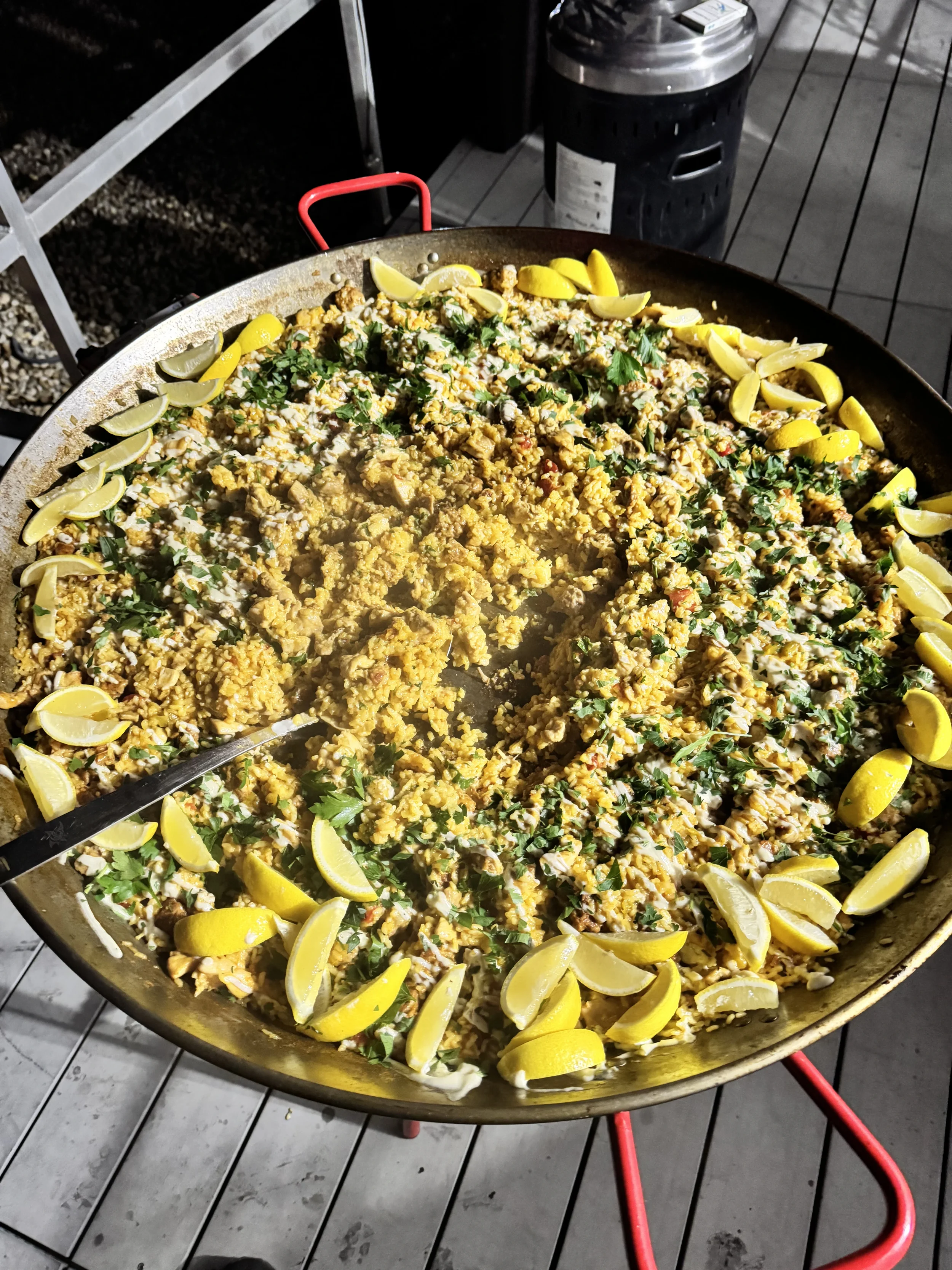 Large outdoor paella cooked in a pan with lemon wedges and herbs, placed on a table at night.