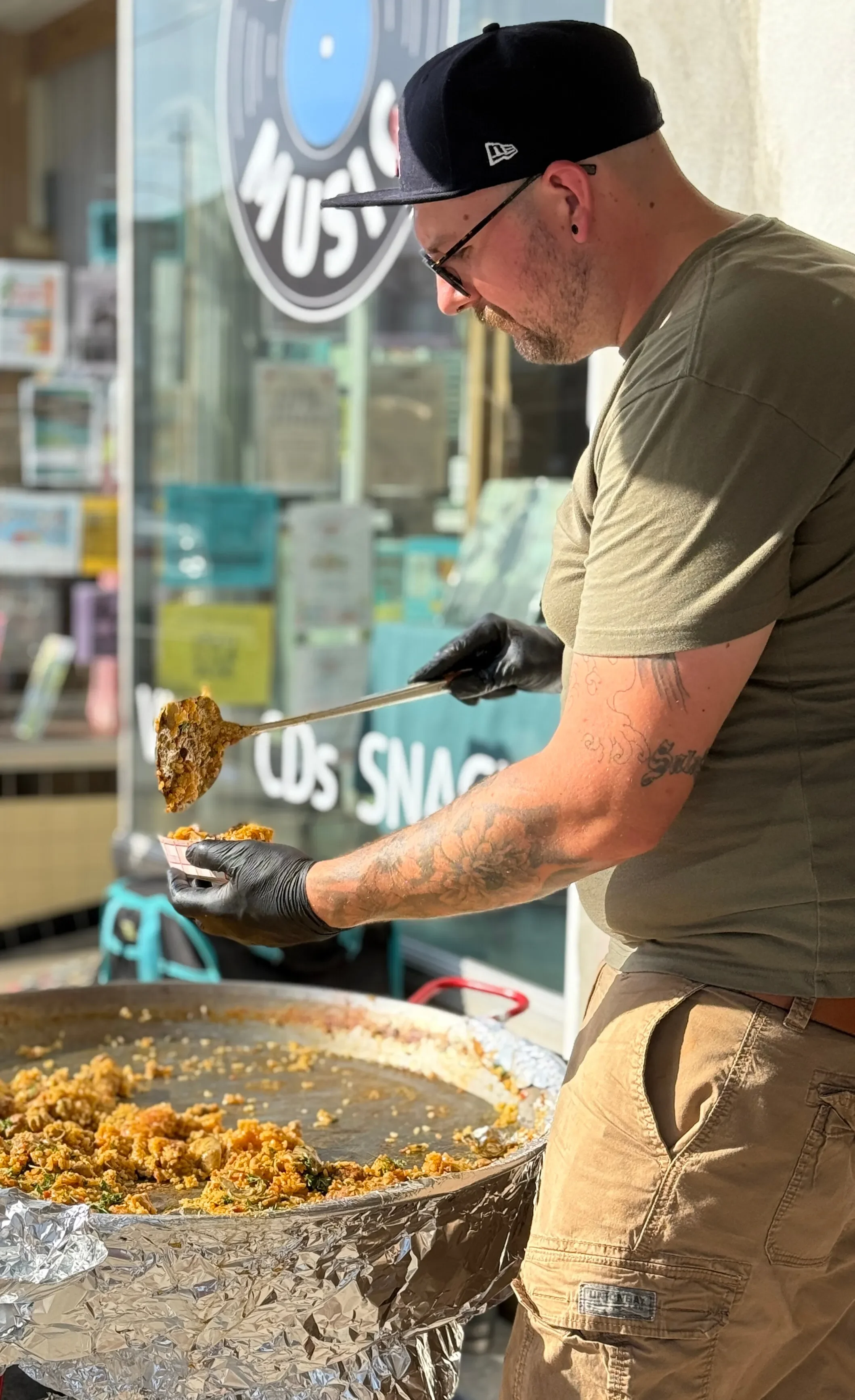 A man wearing a black cap, glasses, a green shirt, and black gloves serving food from a large pan at a food stand. The food appears to be a type of seasoned rice or grain dish. There is a sign in the background that reads 'Los Snacks' and 'Music'.