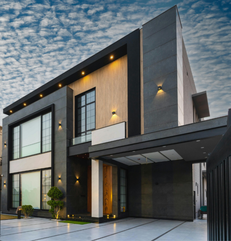 Modern multi-story house with black and gray exterior, large windows, and outdoor lighting, under a partly cloudy sky at dusk.