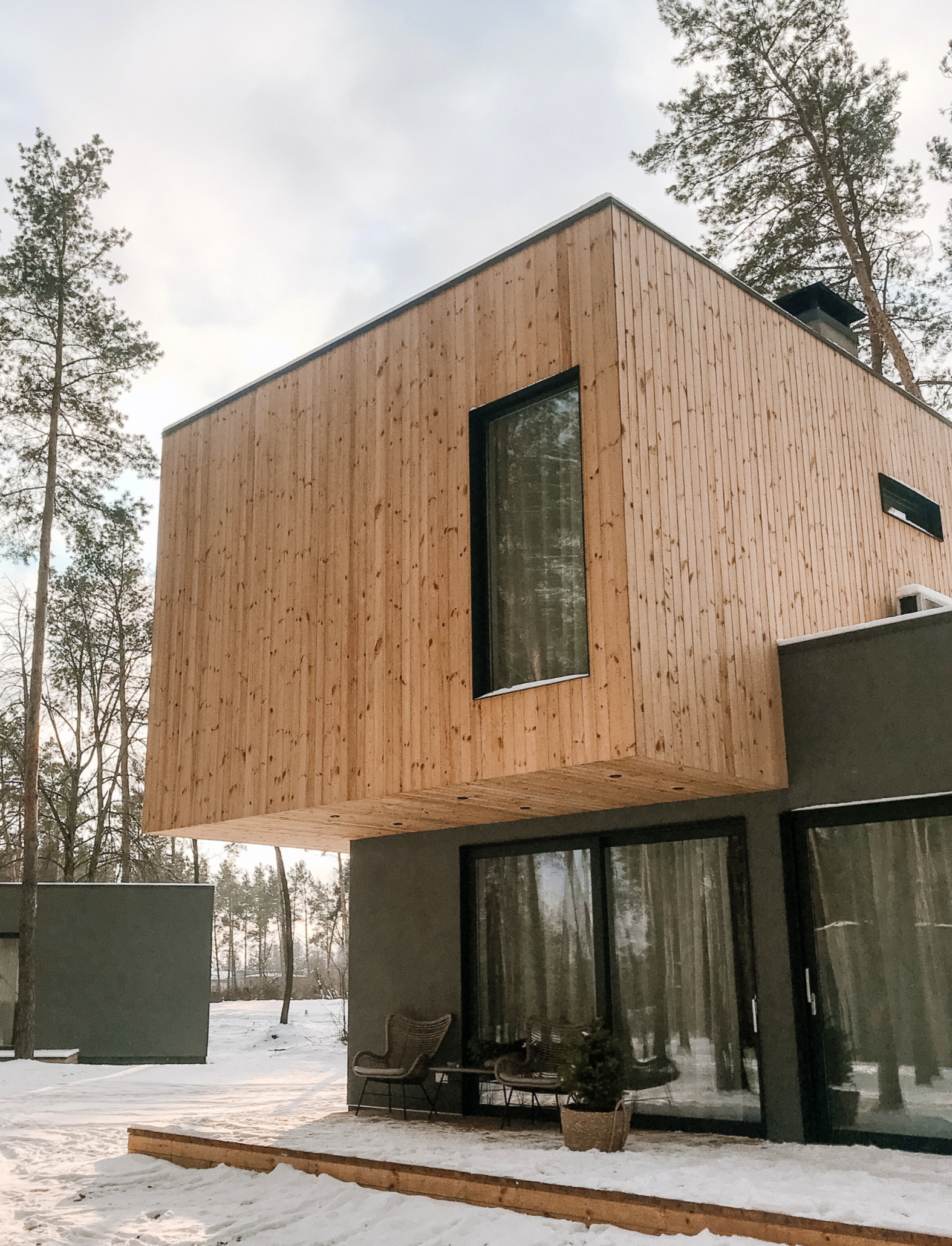 Modern house with wooden and dark exterior, large windows, snow-covered ground, potted plant, outdoor chairs, surrounded by tall trees.