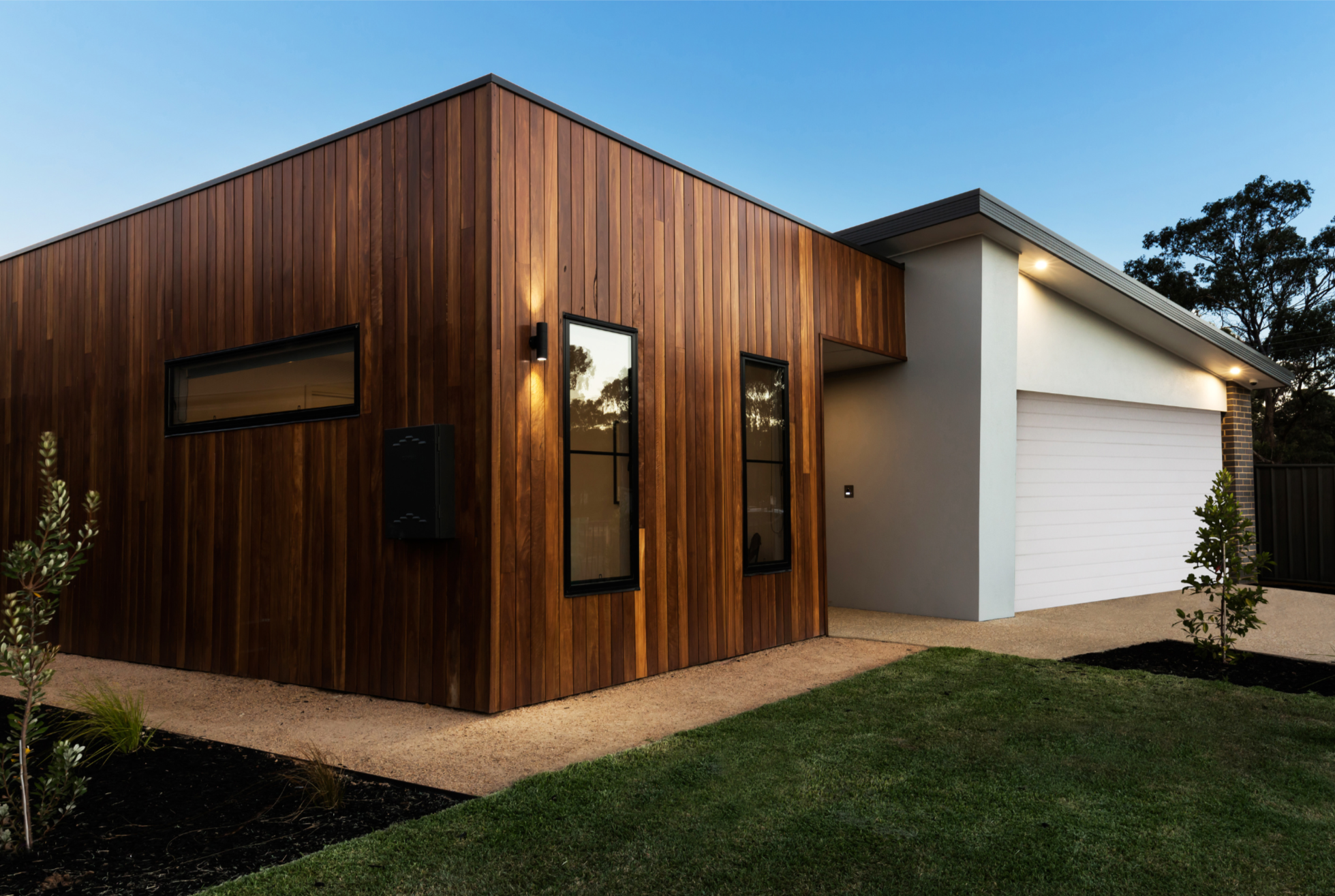 Modern house with wooden and white exterior finishes, featuring two tall narrow windows, a garage door, and exterior lighting at dusk.