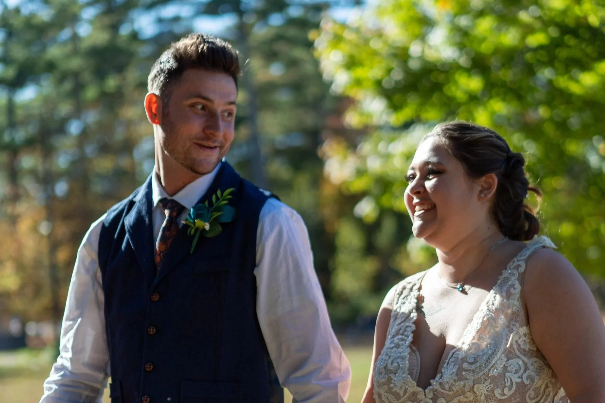 A newlywed couple walking outdoors on a sunny day, holding hands and smiling at each other. The man wears a dark vest over a white shirt with a boutonniere, and the woman wears a lace wedding dress. Trees with green leaves are in the background.