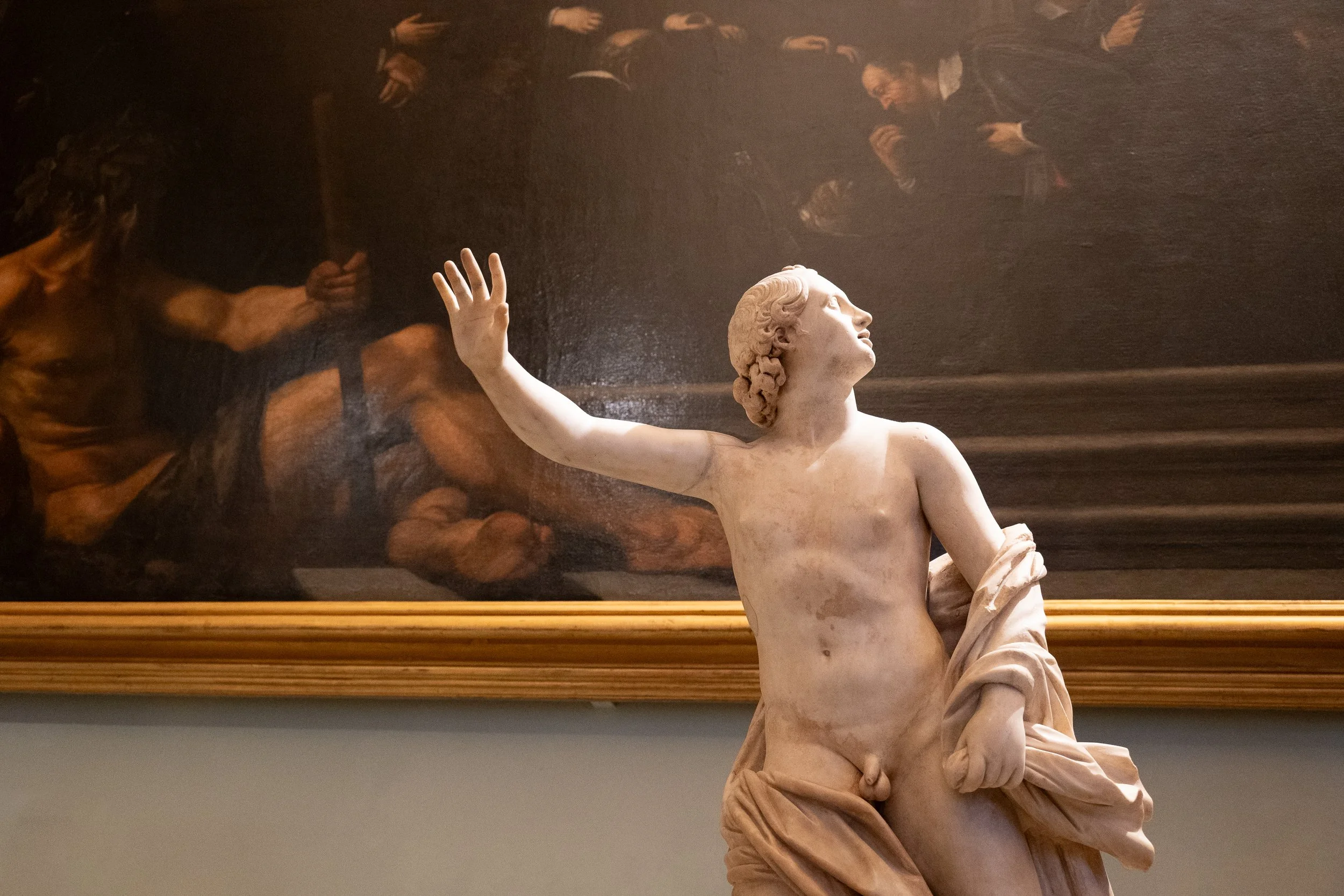 A marble sculpture of a young man with curly hair, holding a draped cloth, in front of a dark painting with several men in suits.