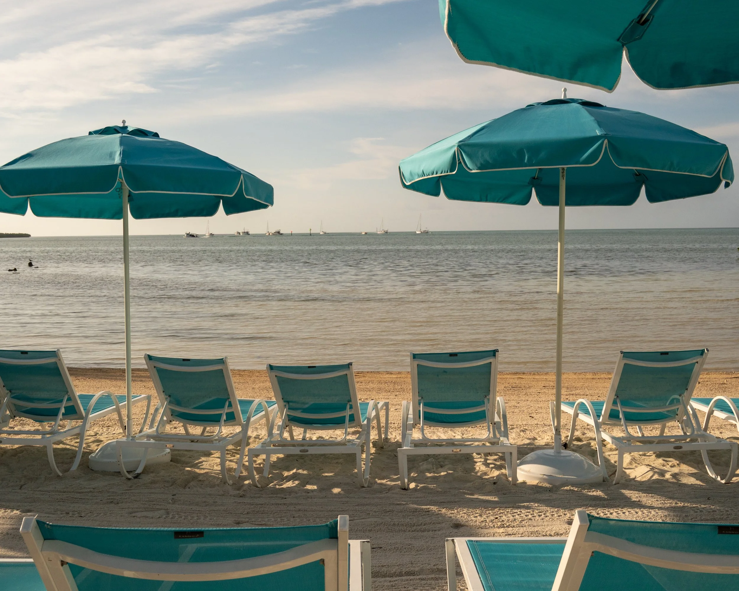 Beach scene with blue umbrellas, lounge chairs, and sailboats on calm water during daytime, Isla Bella Resort, Marathon, Florida Keys, Florida.