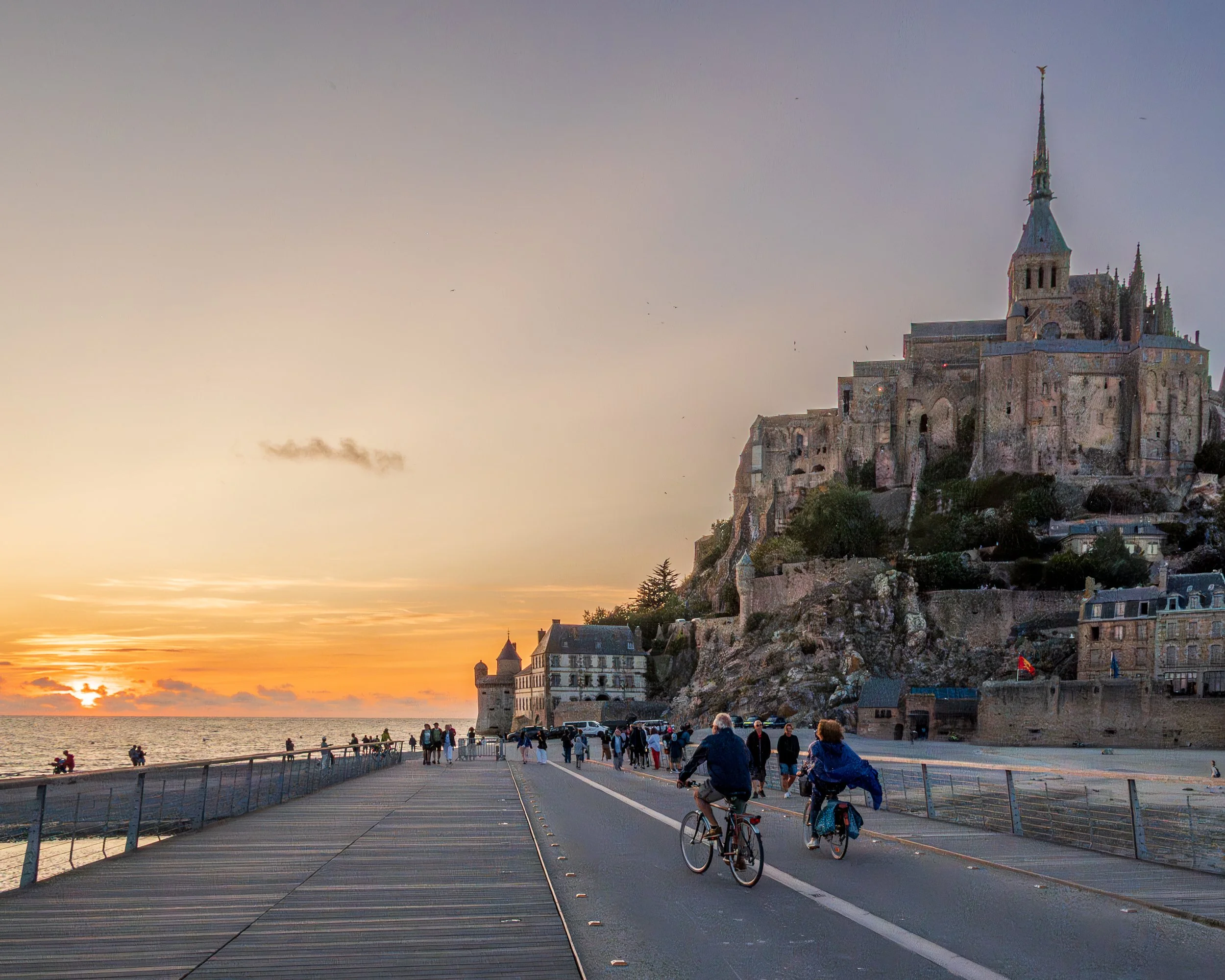 Couple riding bikes next to passersby at sunset, Mont Saint Michel, Normandy region, France.