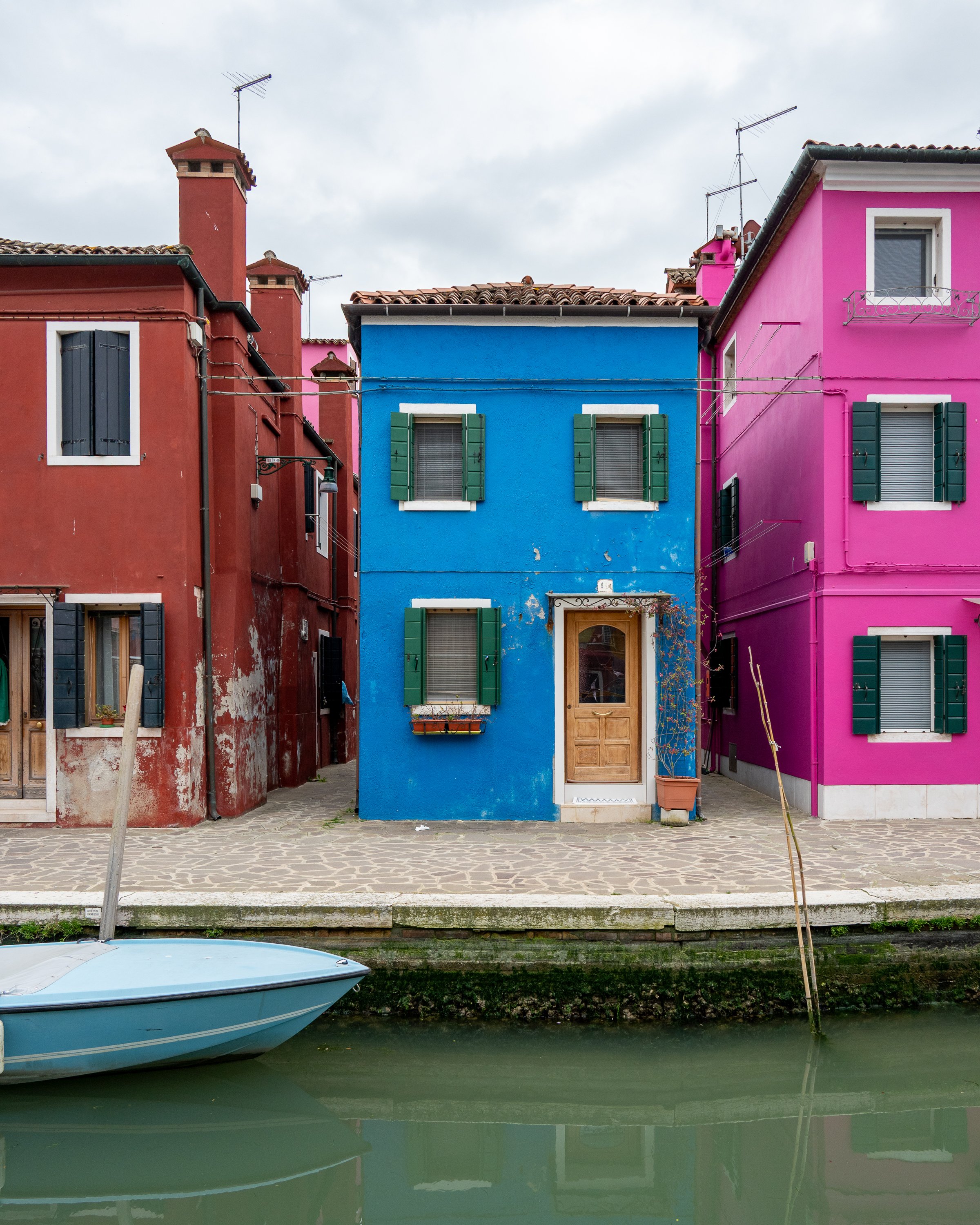Colorful houses along a canal with a boat in the foreground, featuring a blue house in the center with green shutters, flanked by red and pink houses. Burano, Italy.