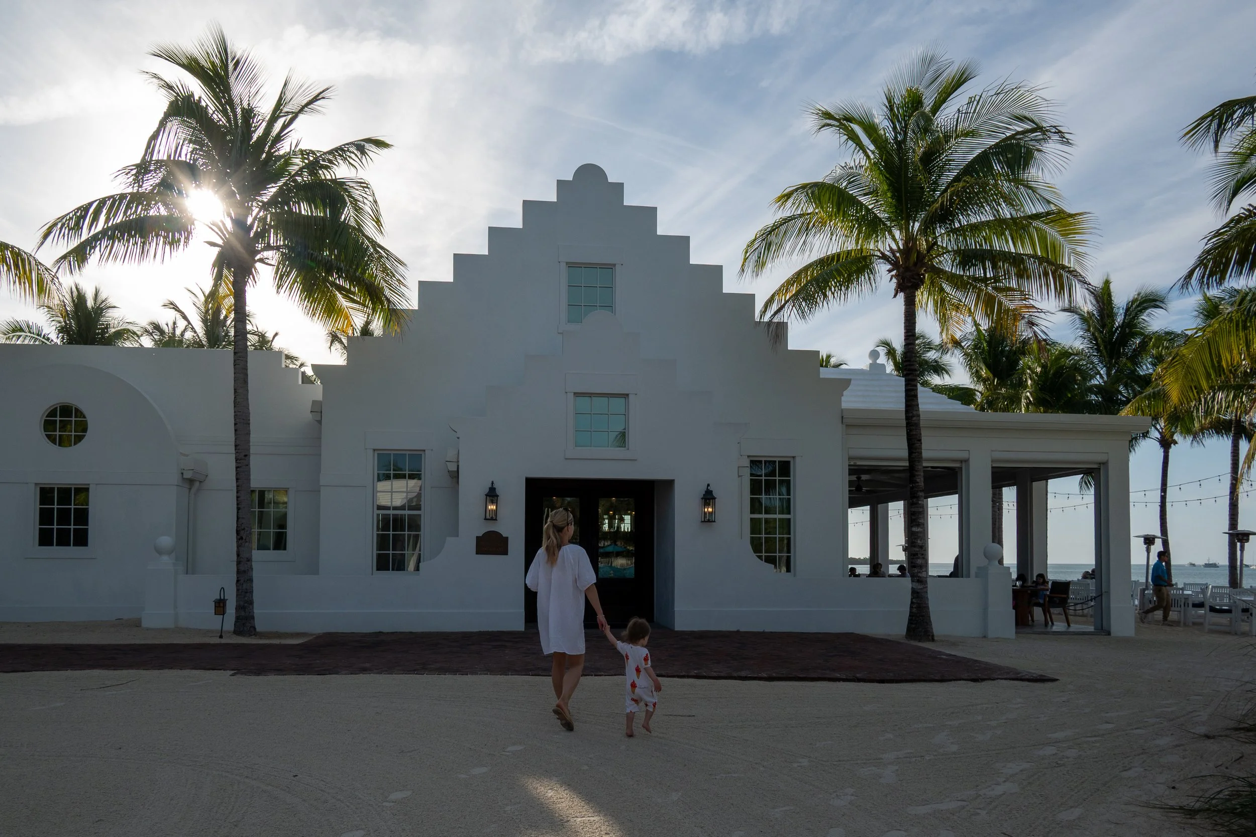 A woman and a child holding hands walking on the sandy beach toward a white building with a stepped gable roof, surrounded by palm trees, with the sun shining through a palm tree on the left, Isla Bella Resort, Marathon, Florida.
