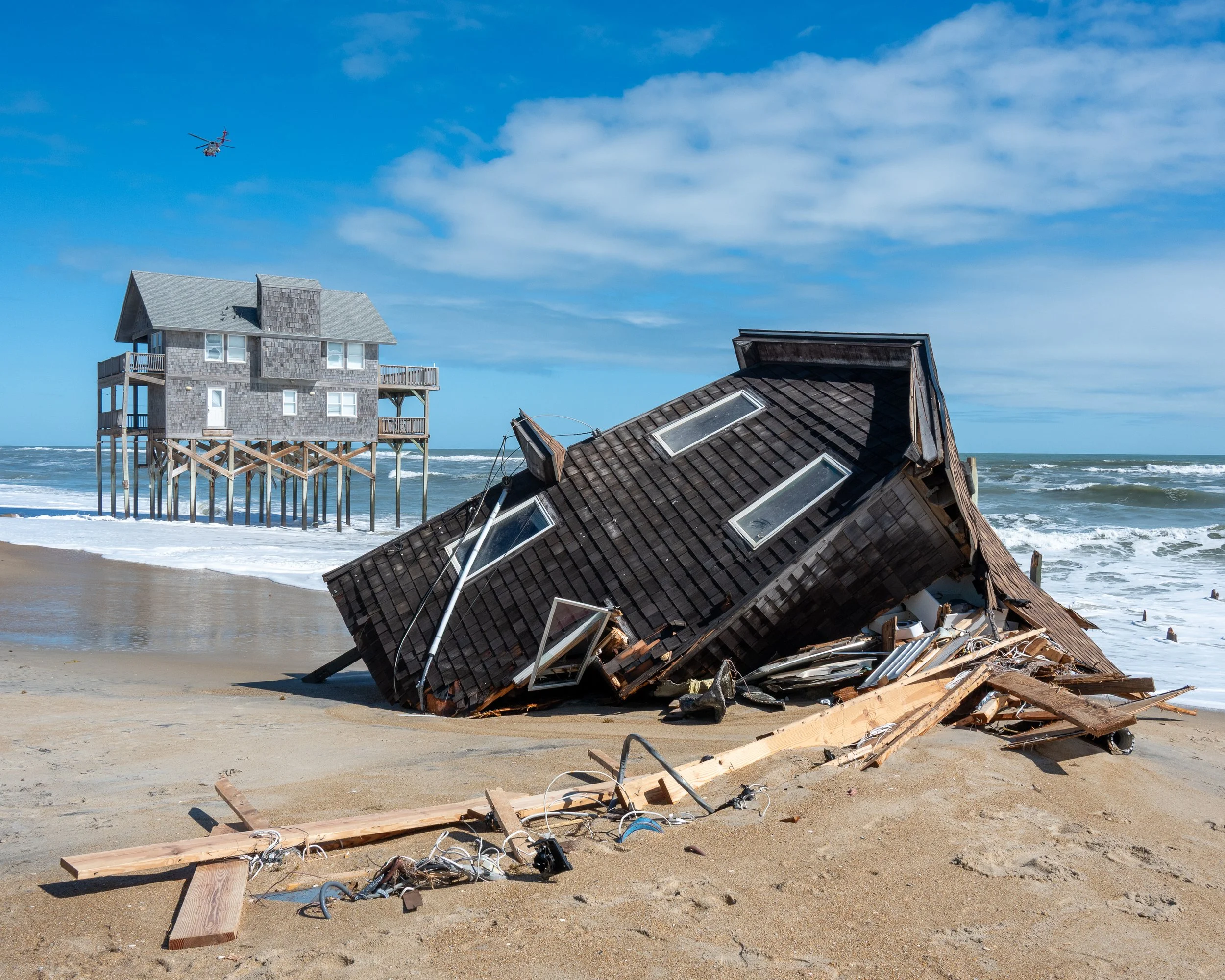 Outer Banks, North Carolina. A collapsed beach house on the sand and another house elevated on stilts in the background, with waves crashing on the shore and a helicopter flying in the sky. 