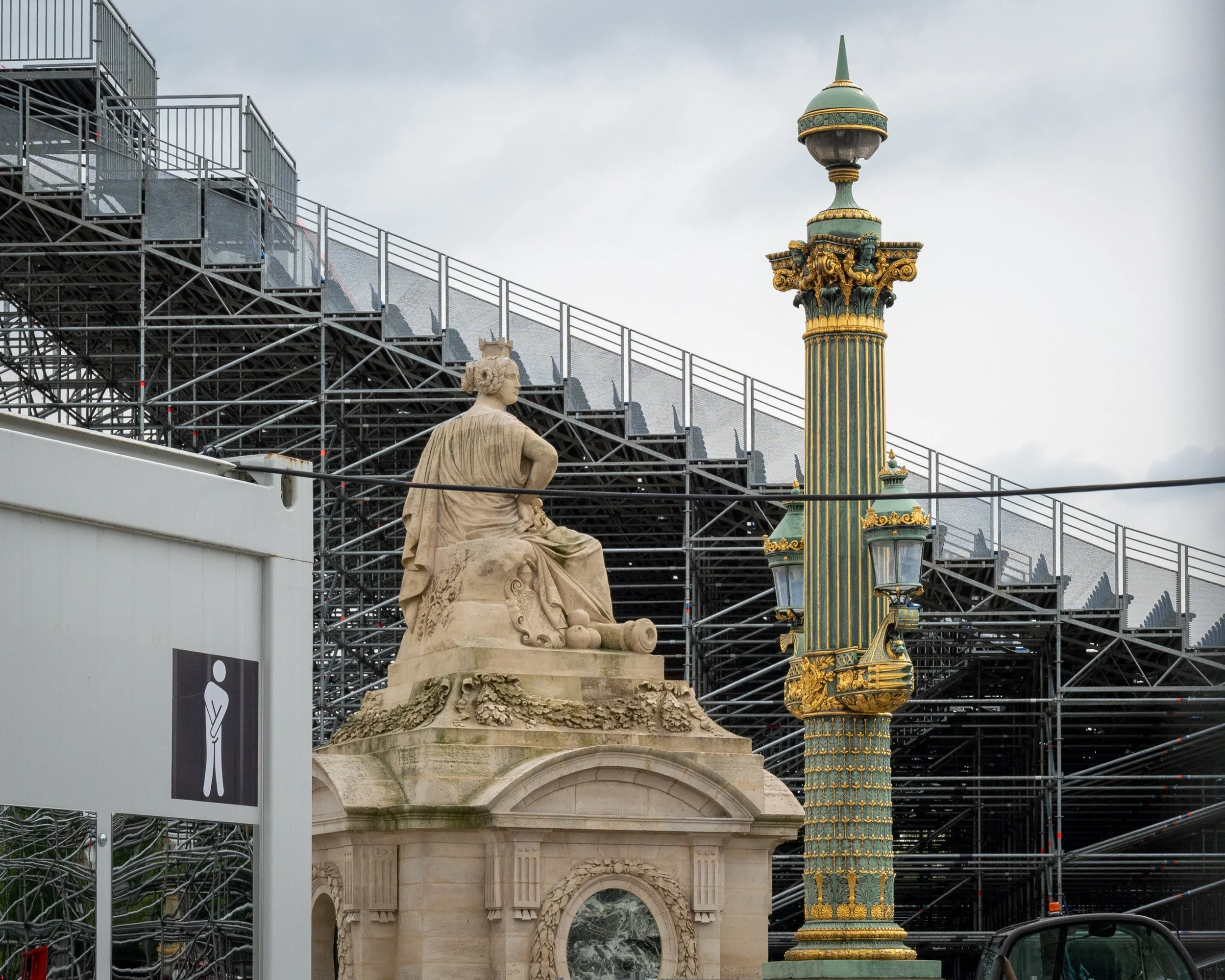 A classical statue of a seated woman and an ornate street lamp with green and gold detailing, in front of a large metal scaffolding structure, ahead of the Paris Olympics, Place de la Concorde, Paris.