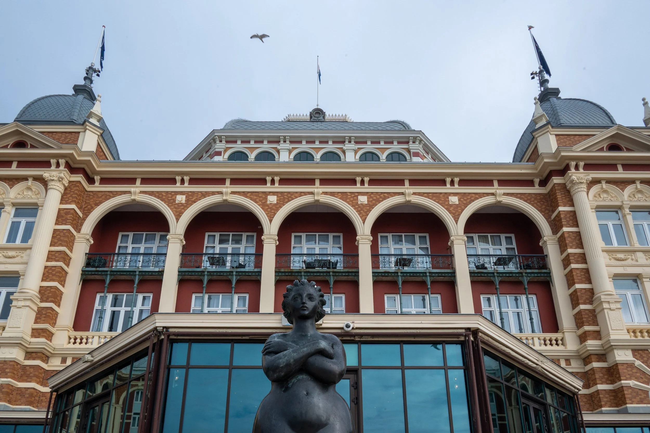 A historic hotel with ornate architecture, multiple balconies, and a statue of a woman in front. The building has a red brick and cream facade with arched windows and decorative columns, The Netherlands.
