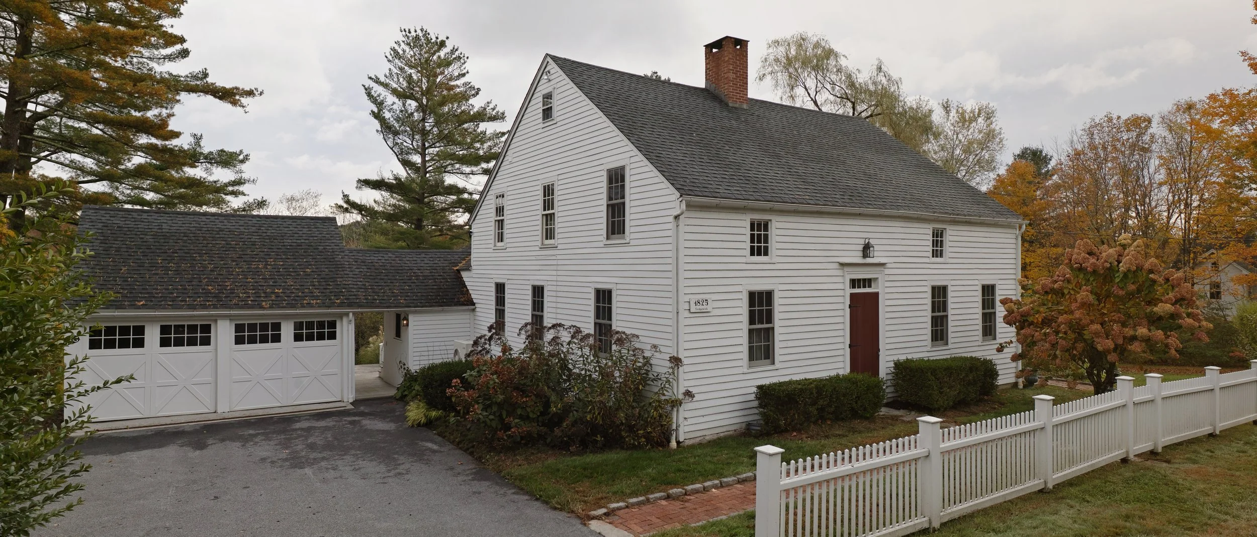 A white two-story house with a gabled roof and red front door, fenced yard, surrounded by trees with autumn leaves, and a white garage with a driveway.