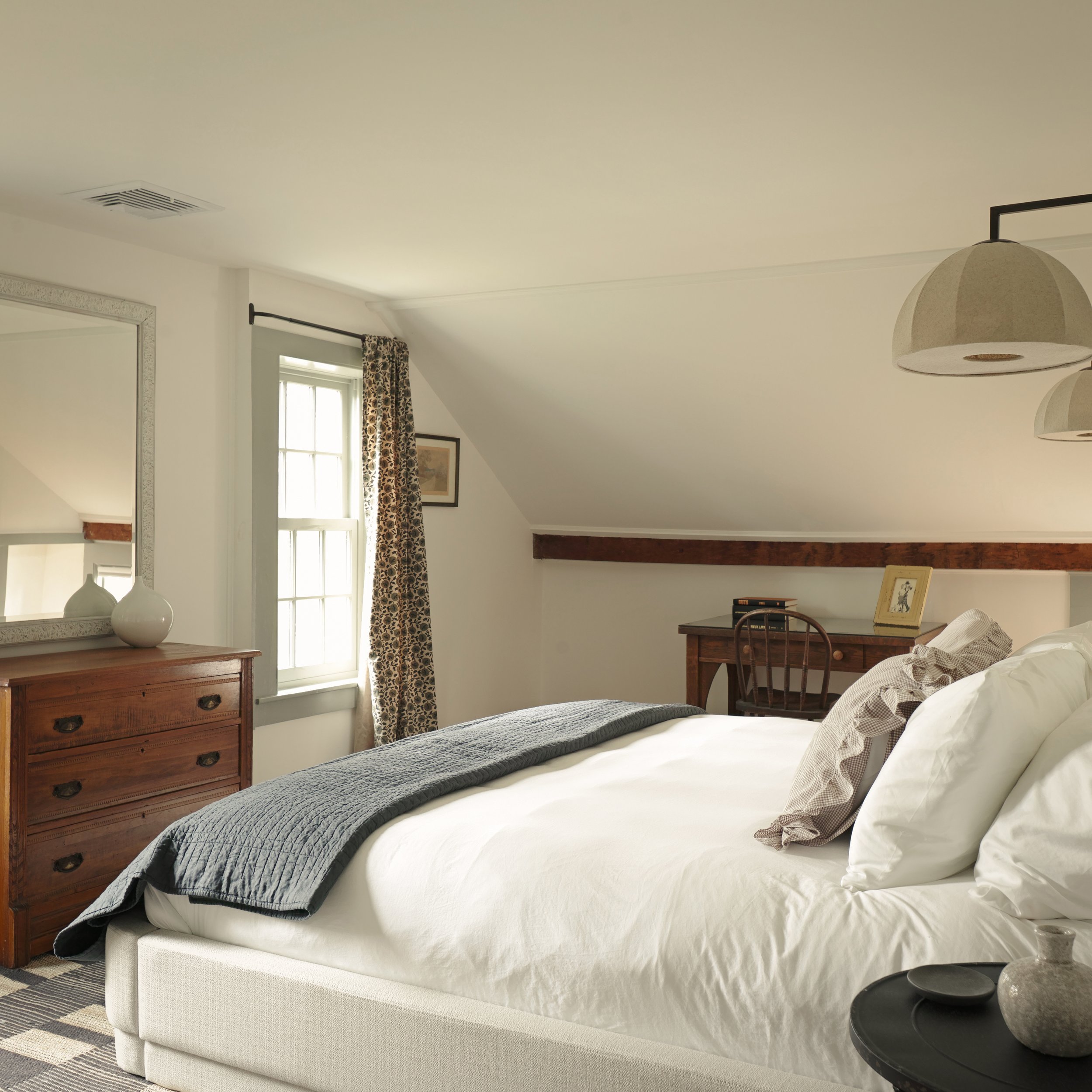 A cozy bedroom with a white bed, gray blanket, and white pillows. Natural light comes through a window with floral curtains. There are wooden furniture pieces, including a dresser, a desk with a chair, and a mirror. Pendant lights hang from the ceili