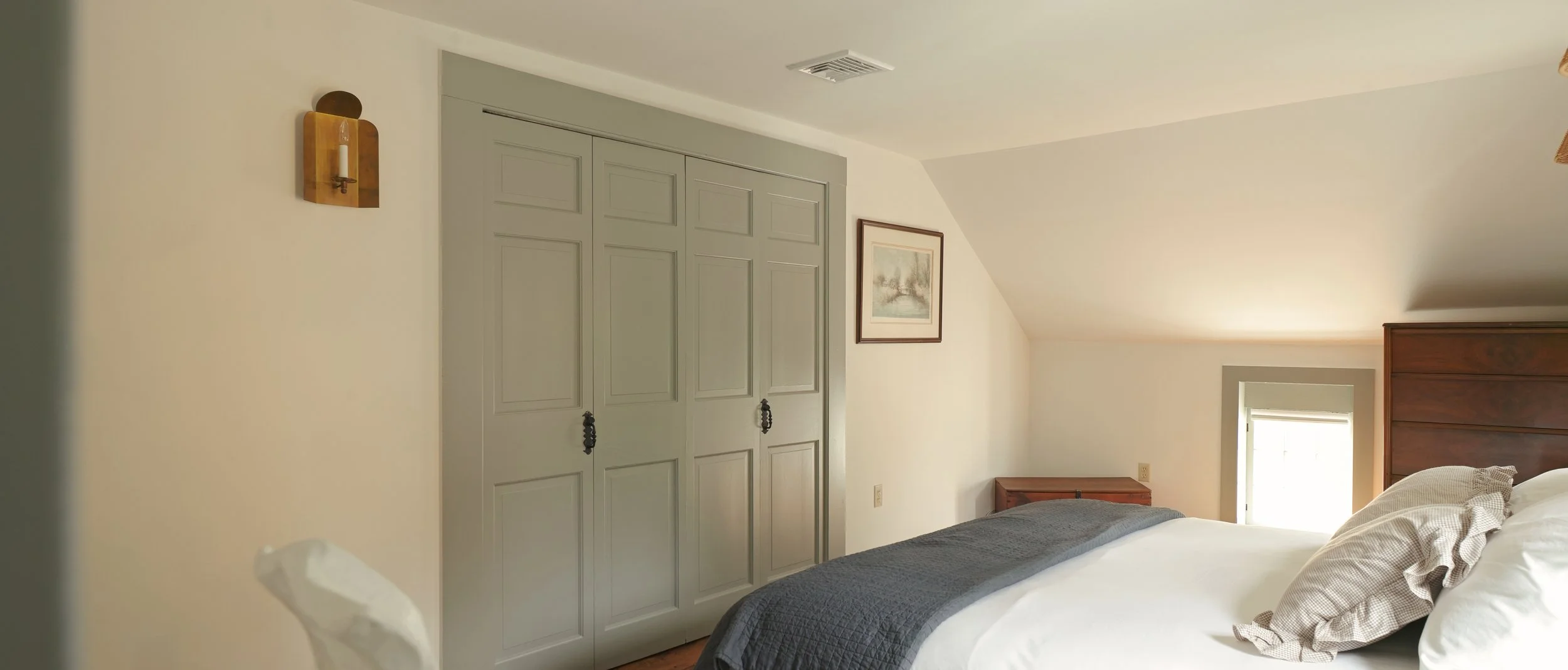 A bedroom with a bed, pillow, blanket, small window, framed picture on the wall, built-in closet, and bedside table.