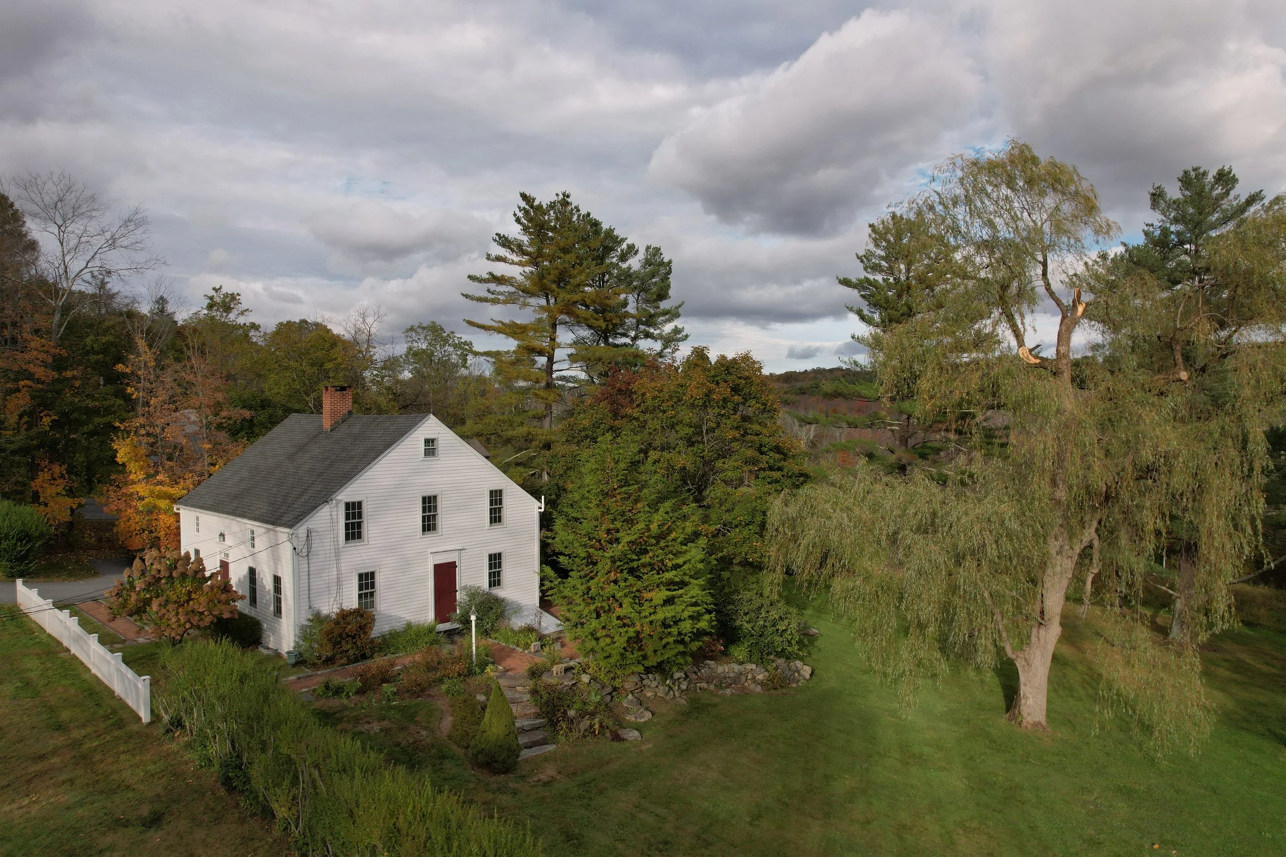 A white house with a gray roof and red door situated in a green yard with tall trees and a cloudy sky.
