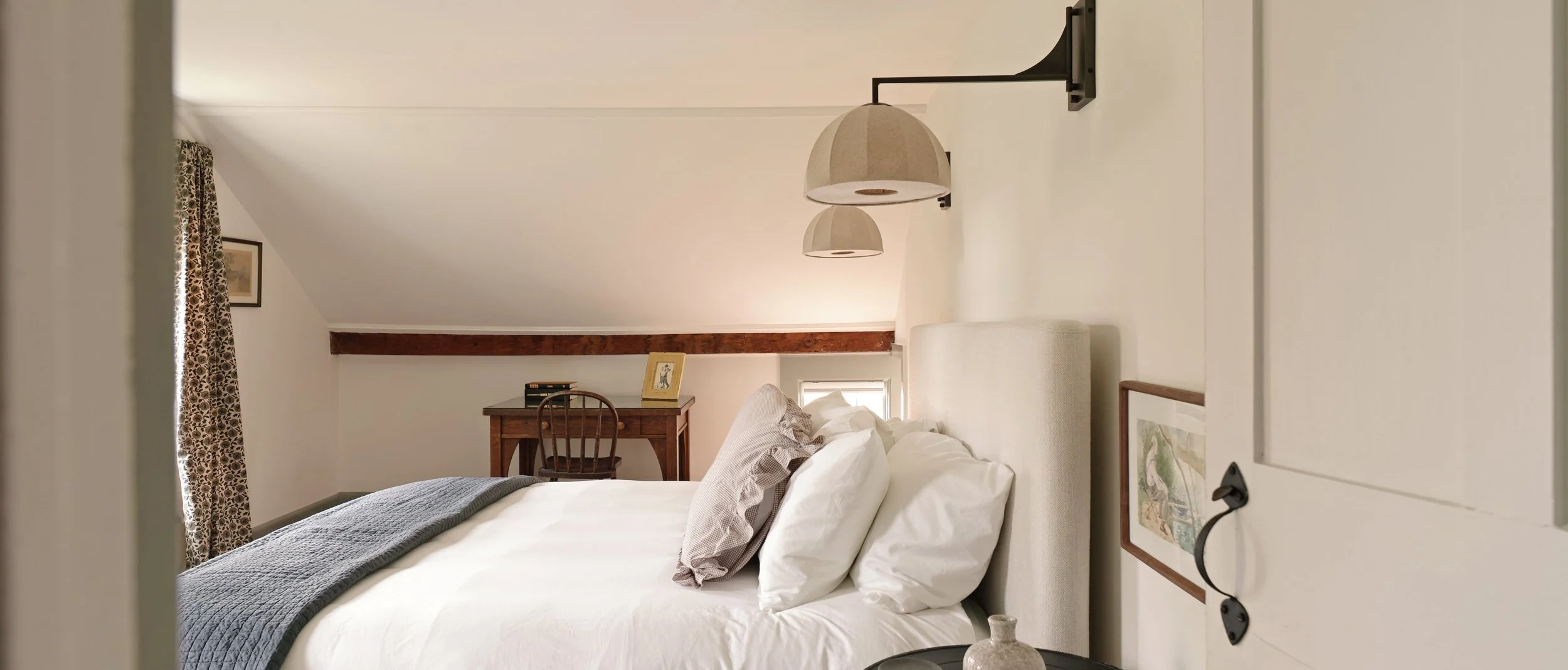A cozy bedroom with a white upholstered bed, gray blanket, multiple pillows, a framed picture on the wall, wooden beams, a small window, and a desk with a chair and books.