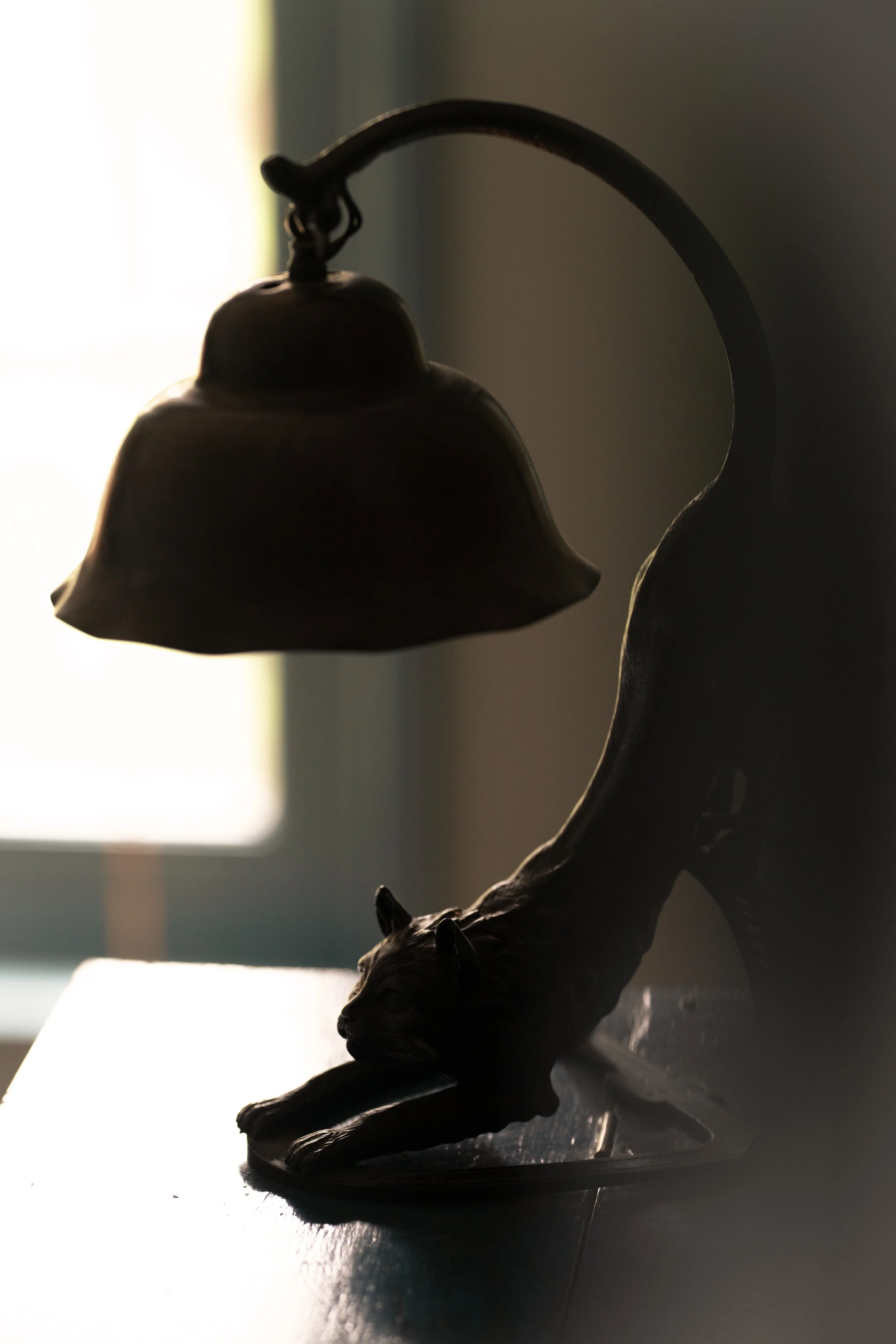 A bronze cat figurine stretching under a lampshade on a table, with light from a window in the background.