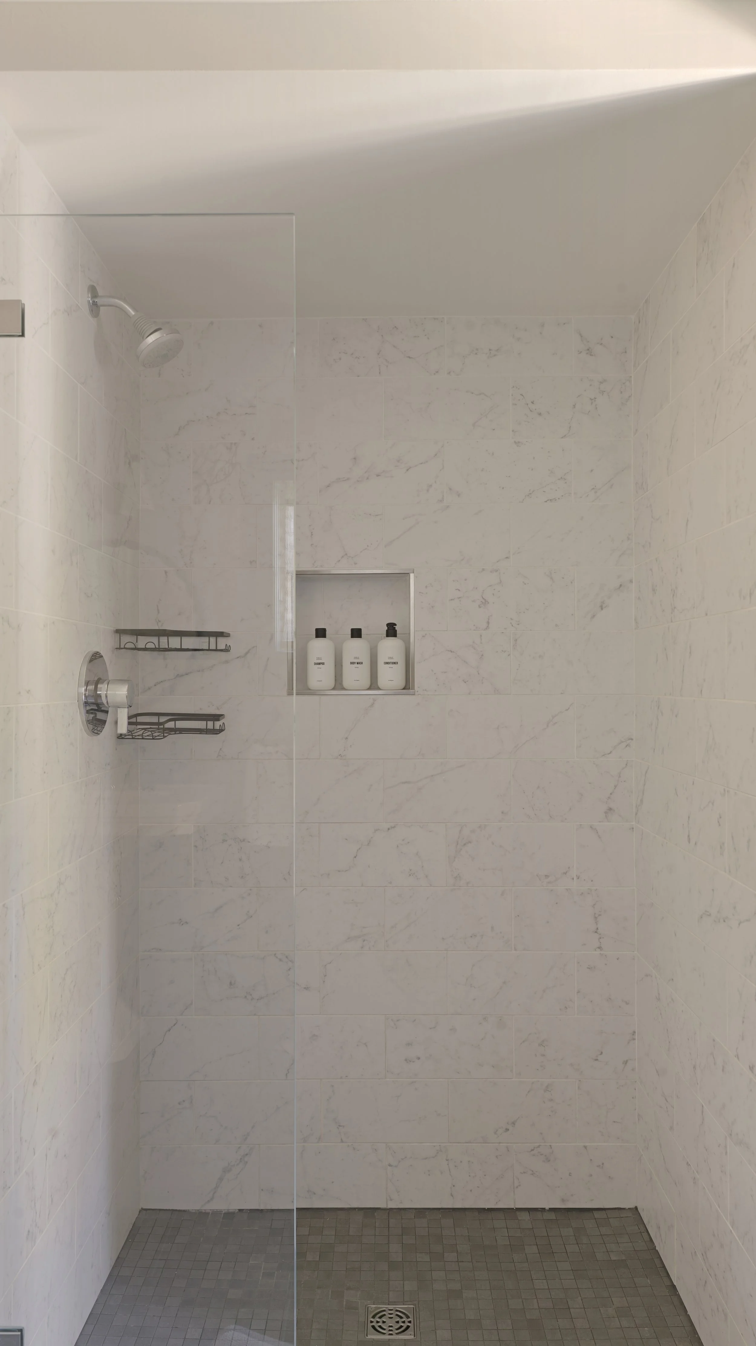 A minimalist shower with white marble tiles, a chrome showerhead, a built-in niche with three bottles, and a glass shower door.