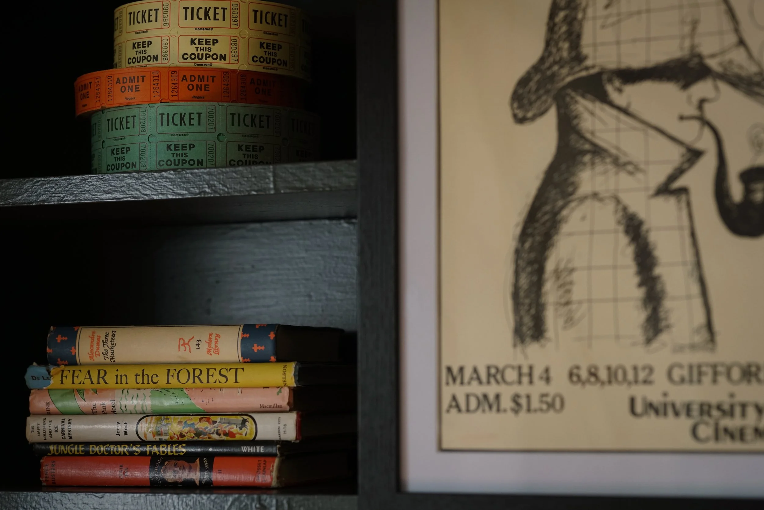 Shelf containing collection of movie tickets in yellow, orange, and green, and a row of books including 'FEAR in the FOREST', 'Jungle Doctor's Fables', and other titles, with a sketch of a woman wearing a hat on a paper on the right side.