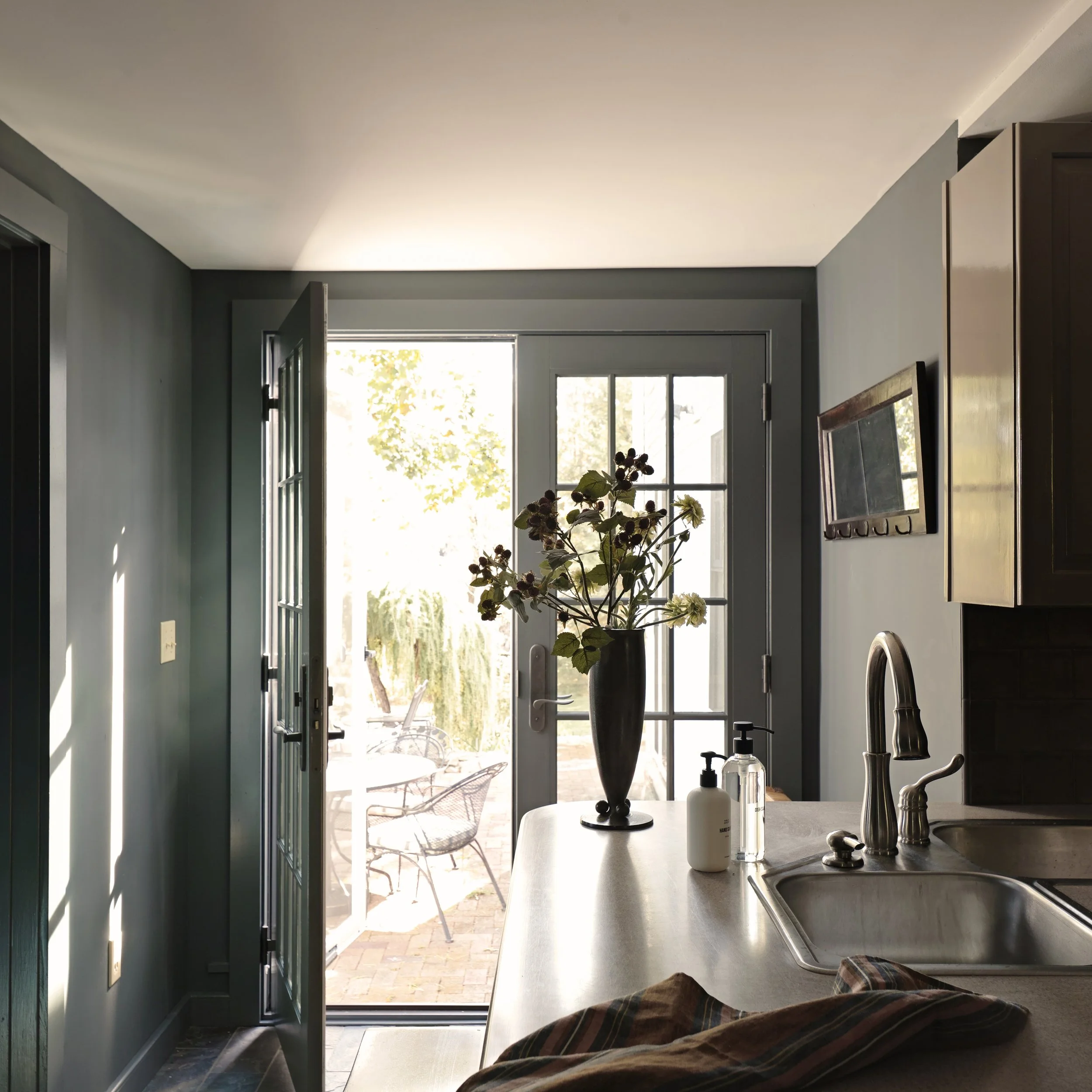 Interior view of a kitchen with an open door leading outside, a countertop with a vase of flowers, soap dispenser, and a faucet, a wall-mounted TV, and a window letting in natural light.