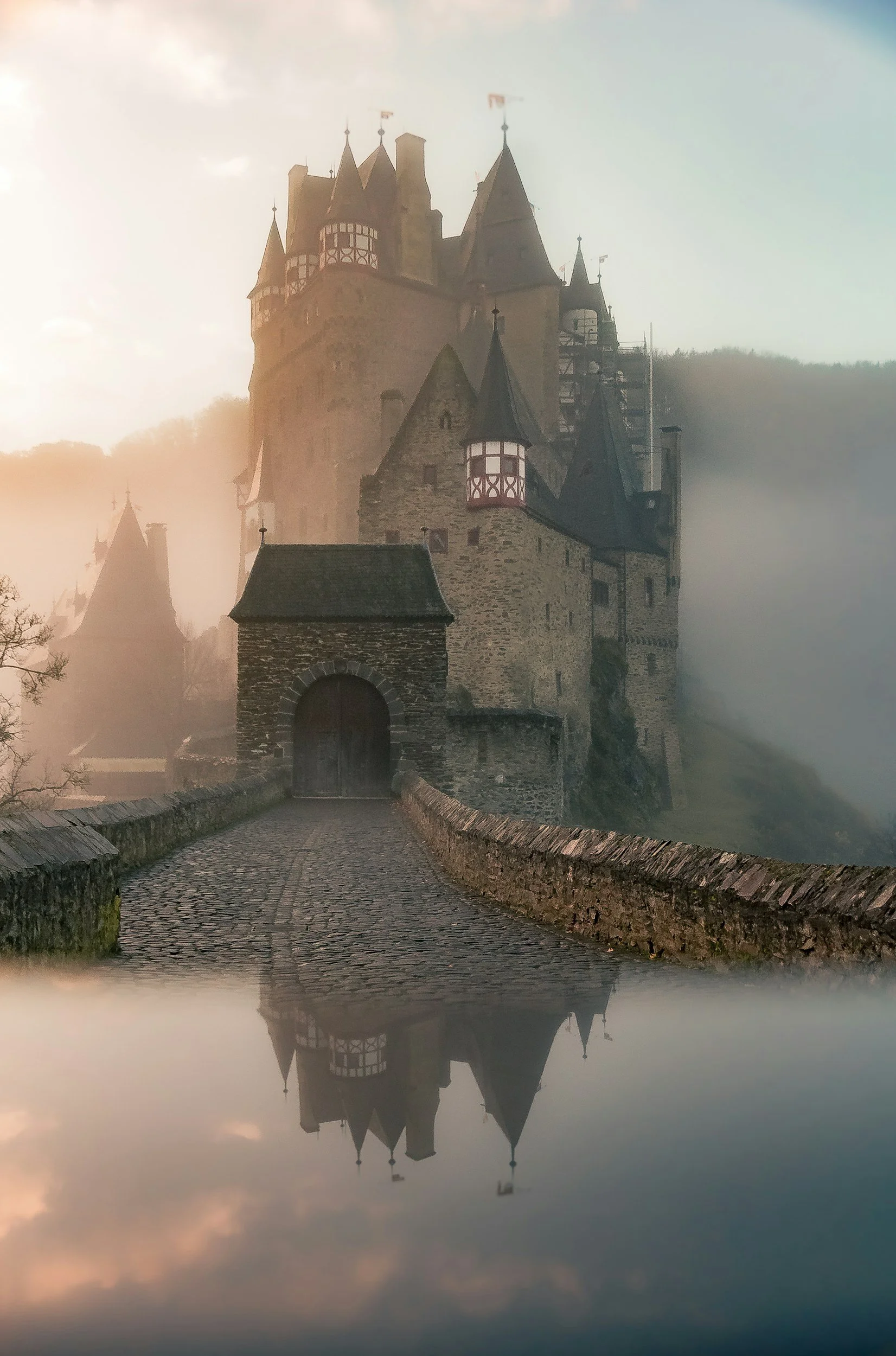A foggy, medieval castle with multiple towers, stone walls, and a cobblestone bridge leading to its entrance, reflected in a body of water.