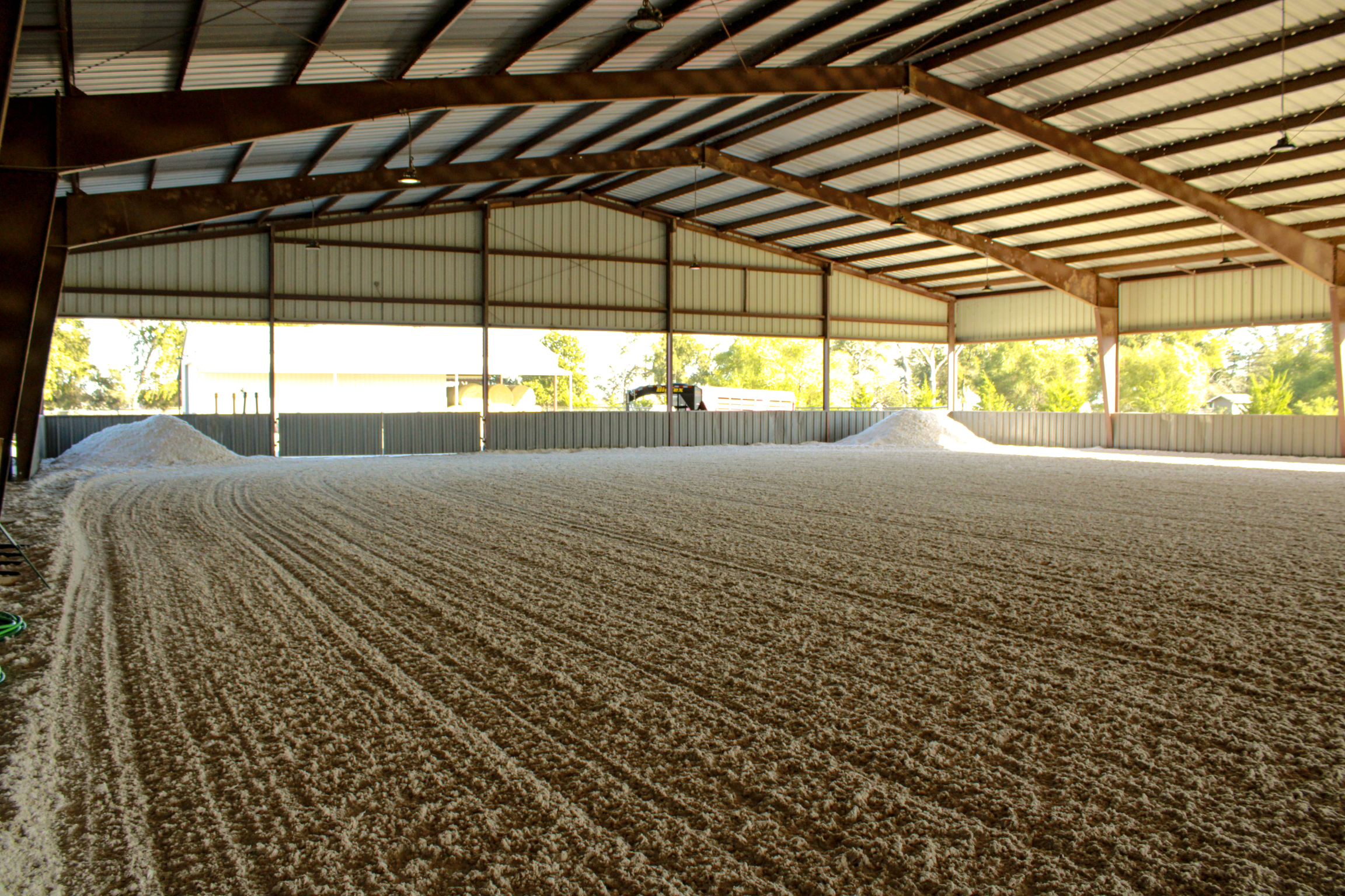 View of an indoor horse riding arena with a sand, metal roof, and exposed metal beams, with some outside greenery visible through open sides.