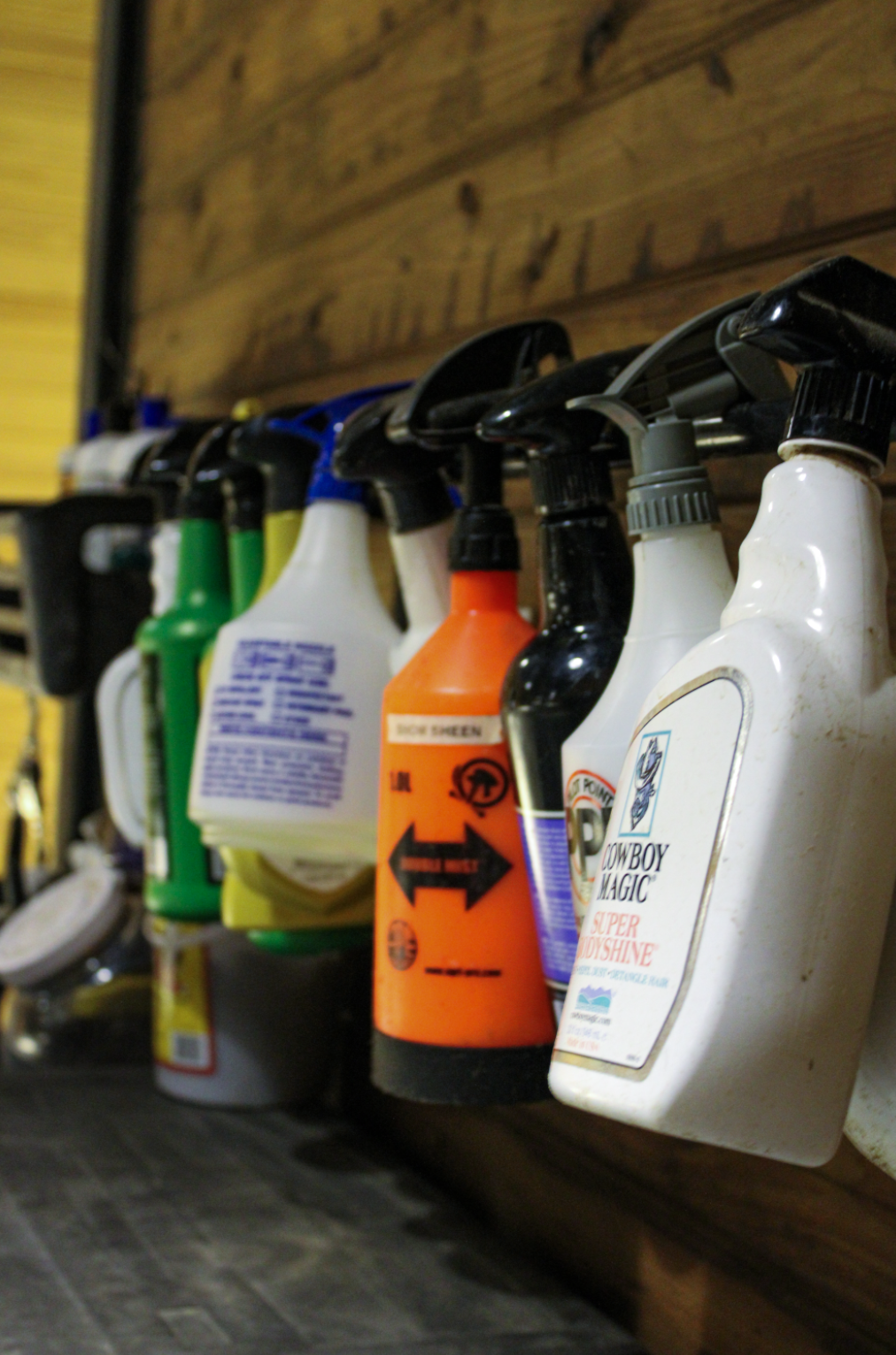 A row of spray bottles and containers hanging on a wooden wall, with some labels visible, including a white bottle labeled 'Cowboy Magic Super Bodyshine' and an orange spray bottle.