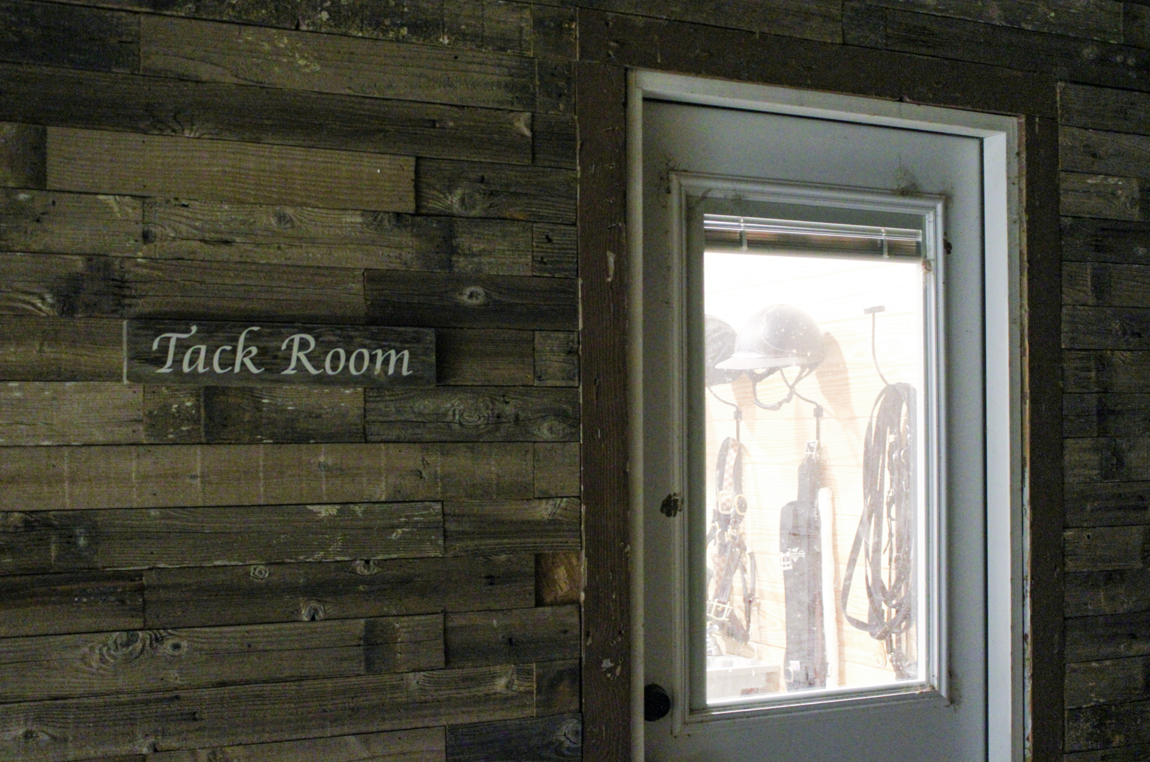 A rustic wooden wall with a small sign that reads 'Tack Room' next to a glass door showing riding helmets and horse tack hanging inside.