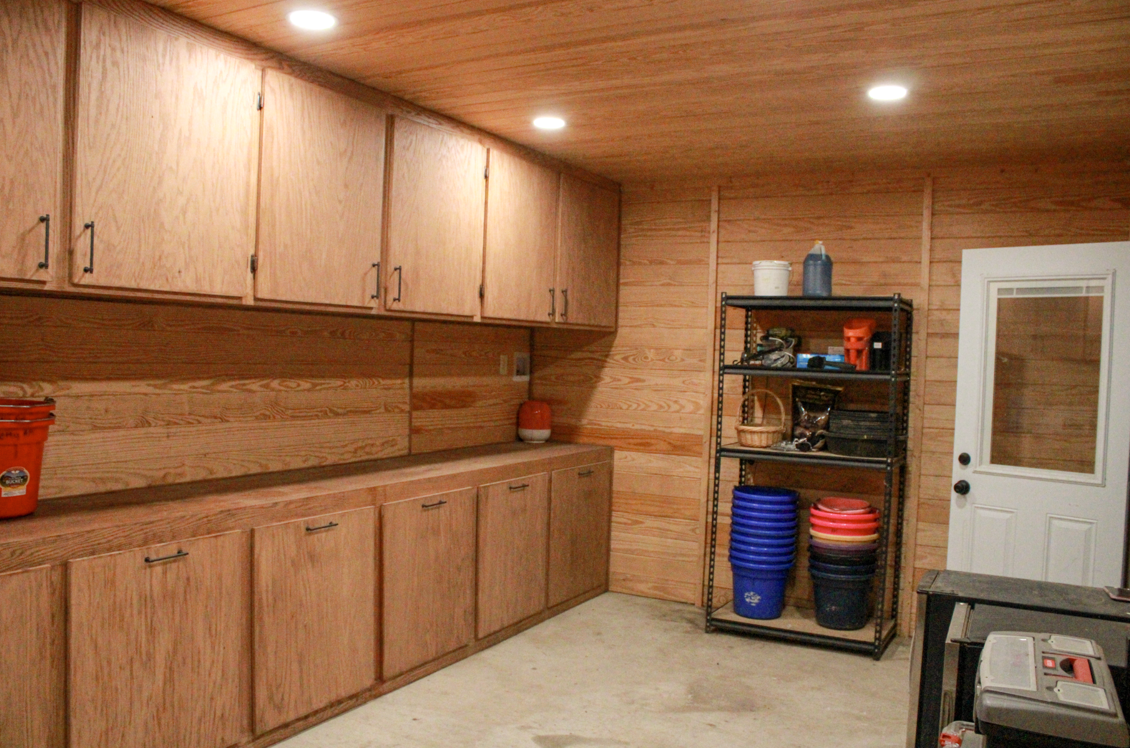 Wood-paneled garage with black metal shelving containing buckets, baskets, and tools, white door with glass window.