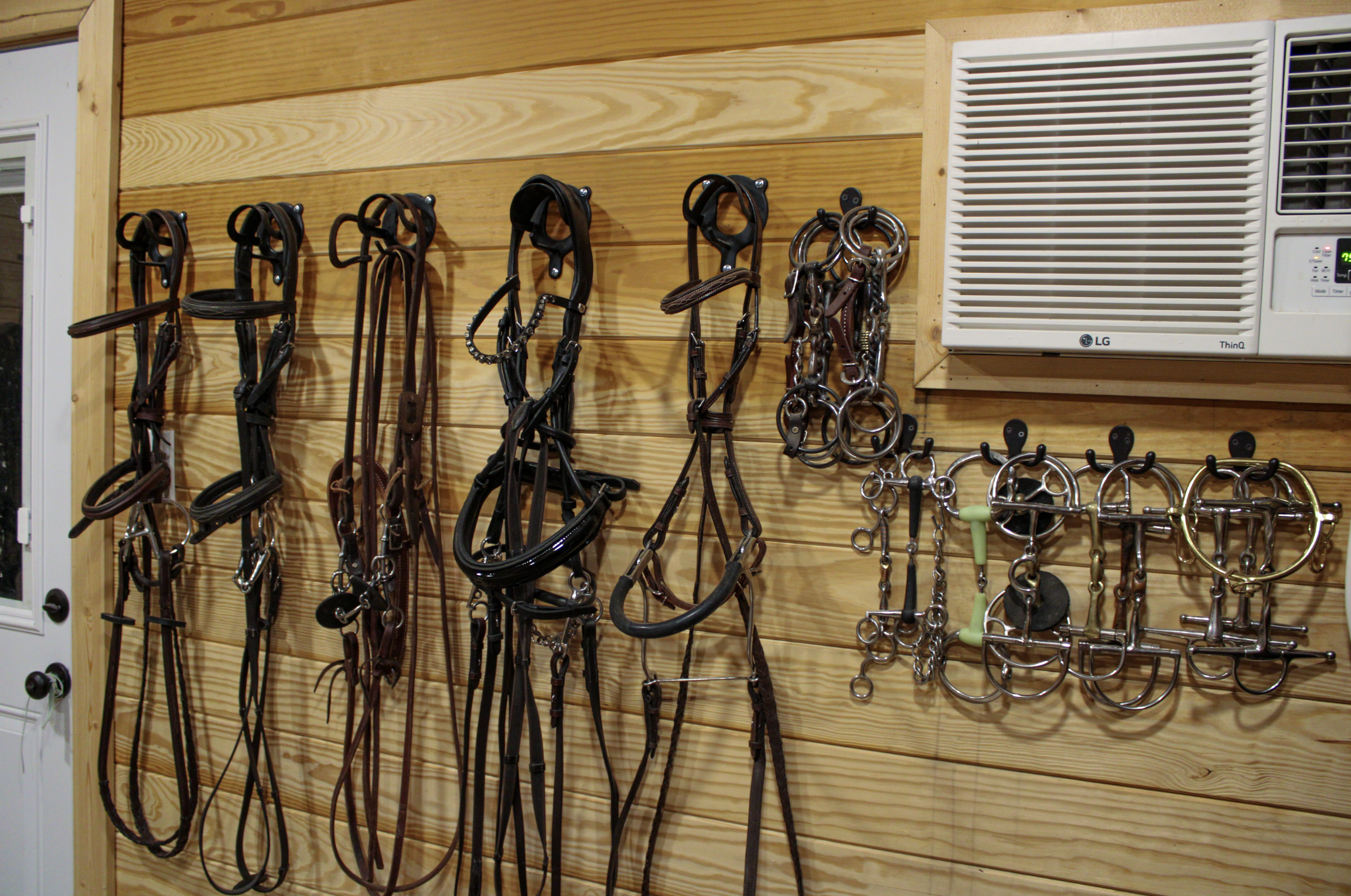 A wall with hanging horse bridles, bits, and bridles on hooks, next to an air conditioning unit. The wall is made of wooden planks.