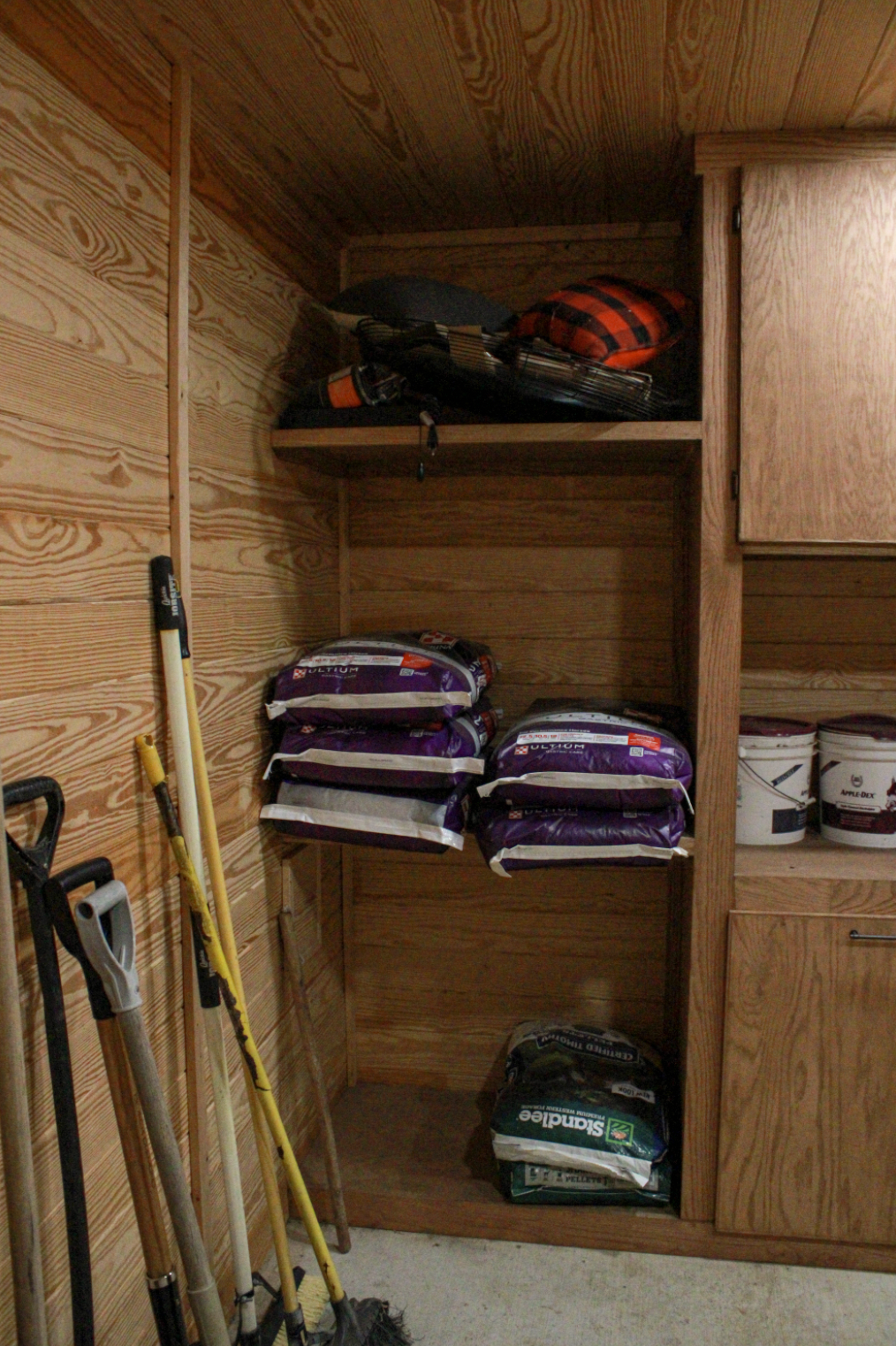 Corner of a wooden storage room with shelves holding bags of horse food, barn supplies, and feed buckets. Rakes and shovels leaning against the wall.
