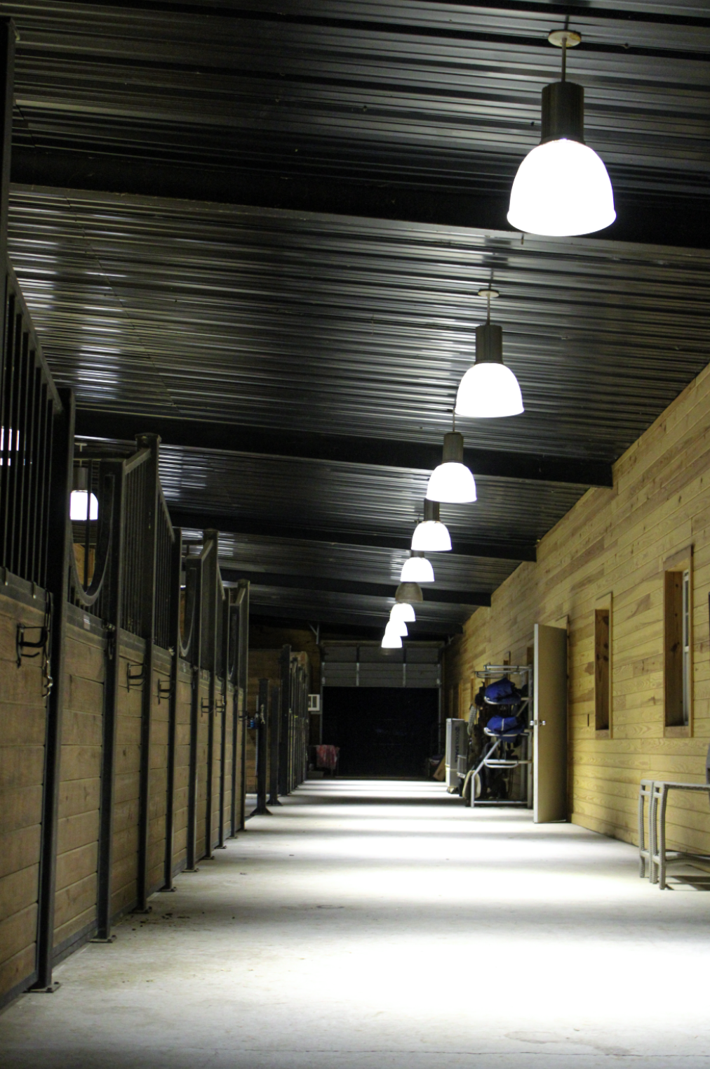 An indoor horse stable with wooden walls, black metal stalls, and bright overhead lights.