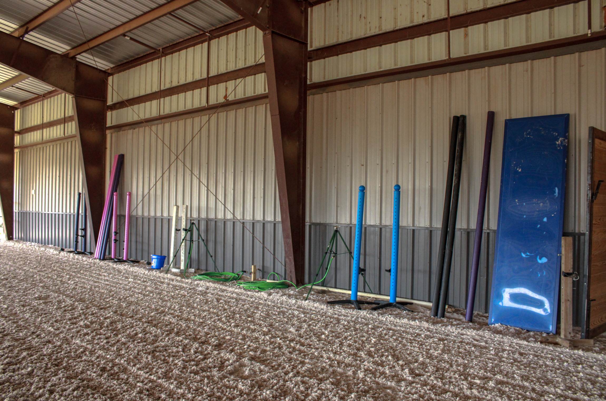 Interior of a metal arena with various poles, a liverpool, and a bucket along the wall, with sand.