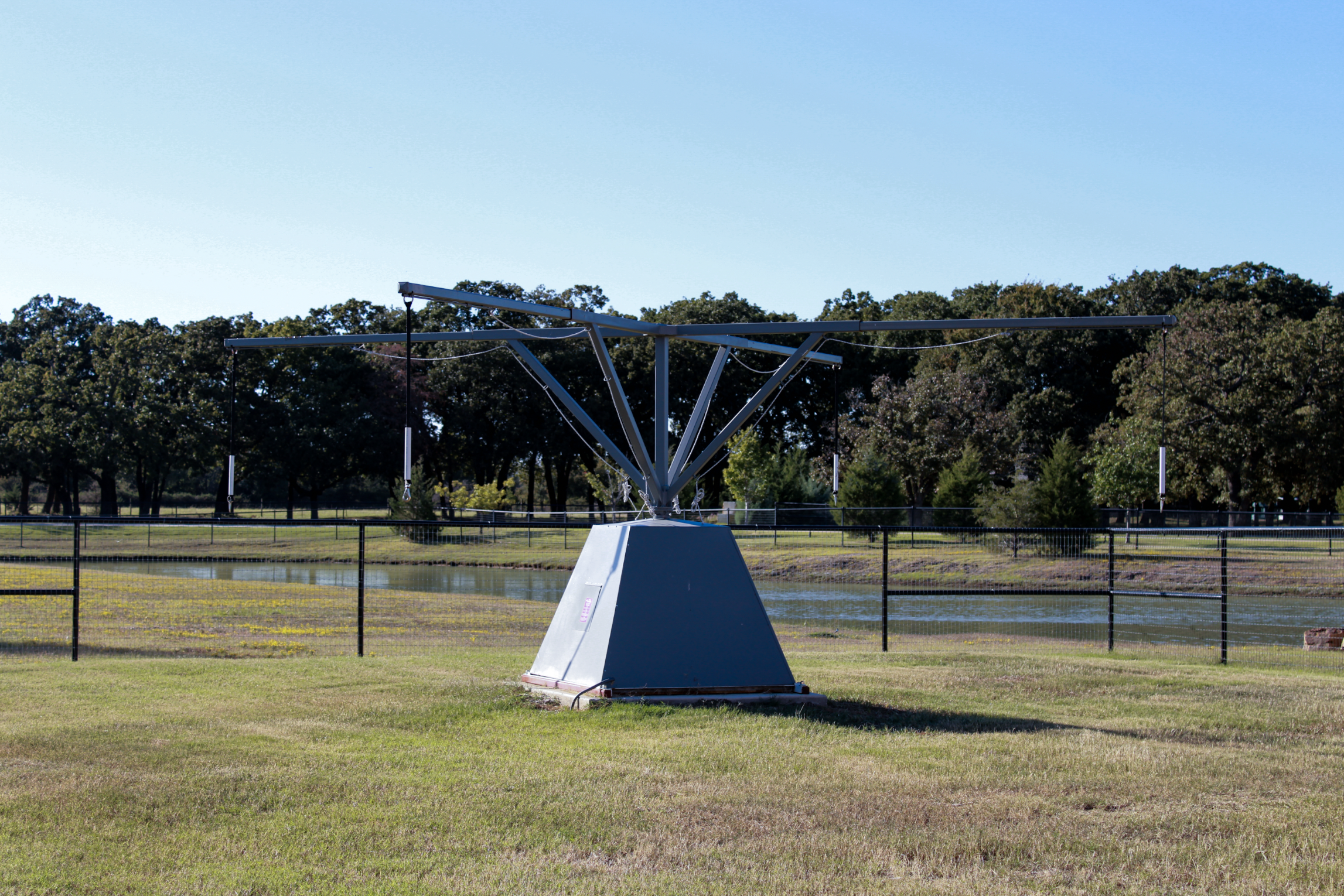 A large metal 4-horse walker situated on a grassy area near a water body with trees in the background.