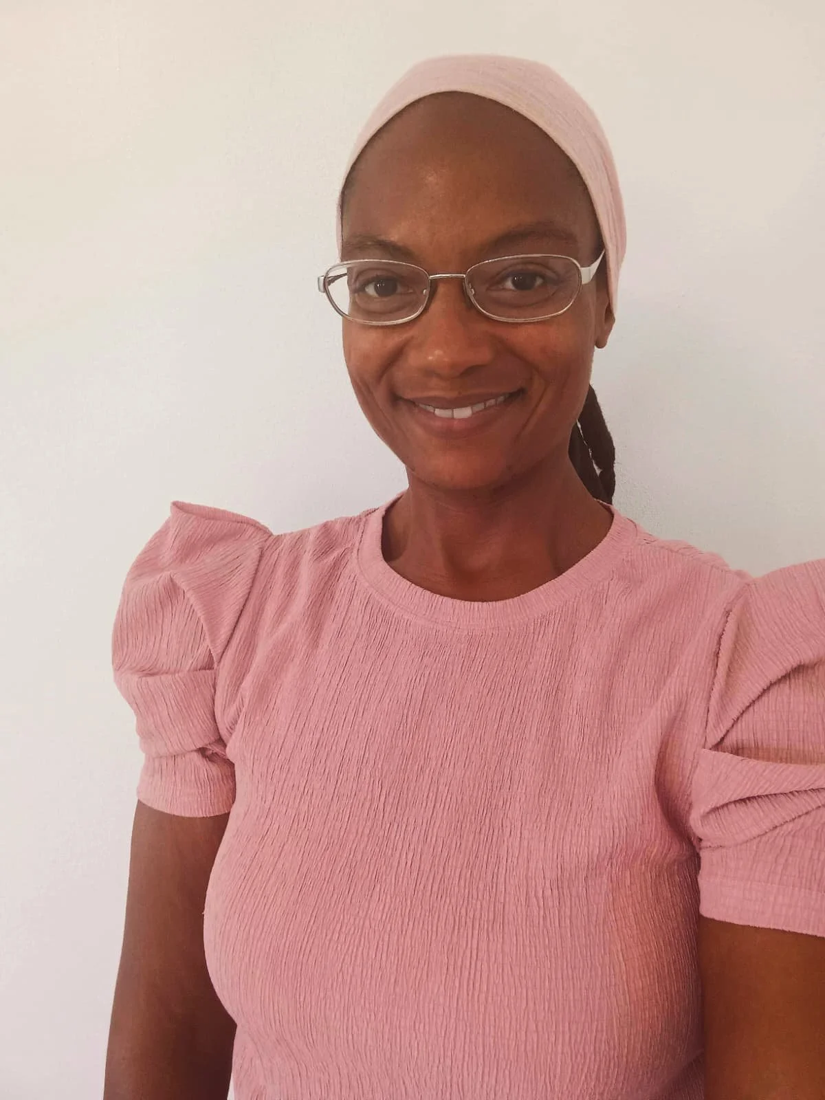 A woman wearing glasses, a yellow headscarf, and a pink jacket in front of green bushes, taking a selfie outdoors.