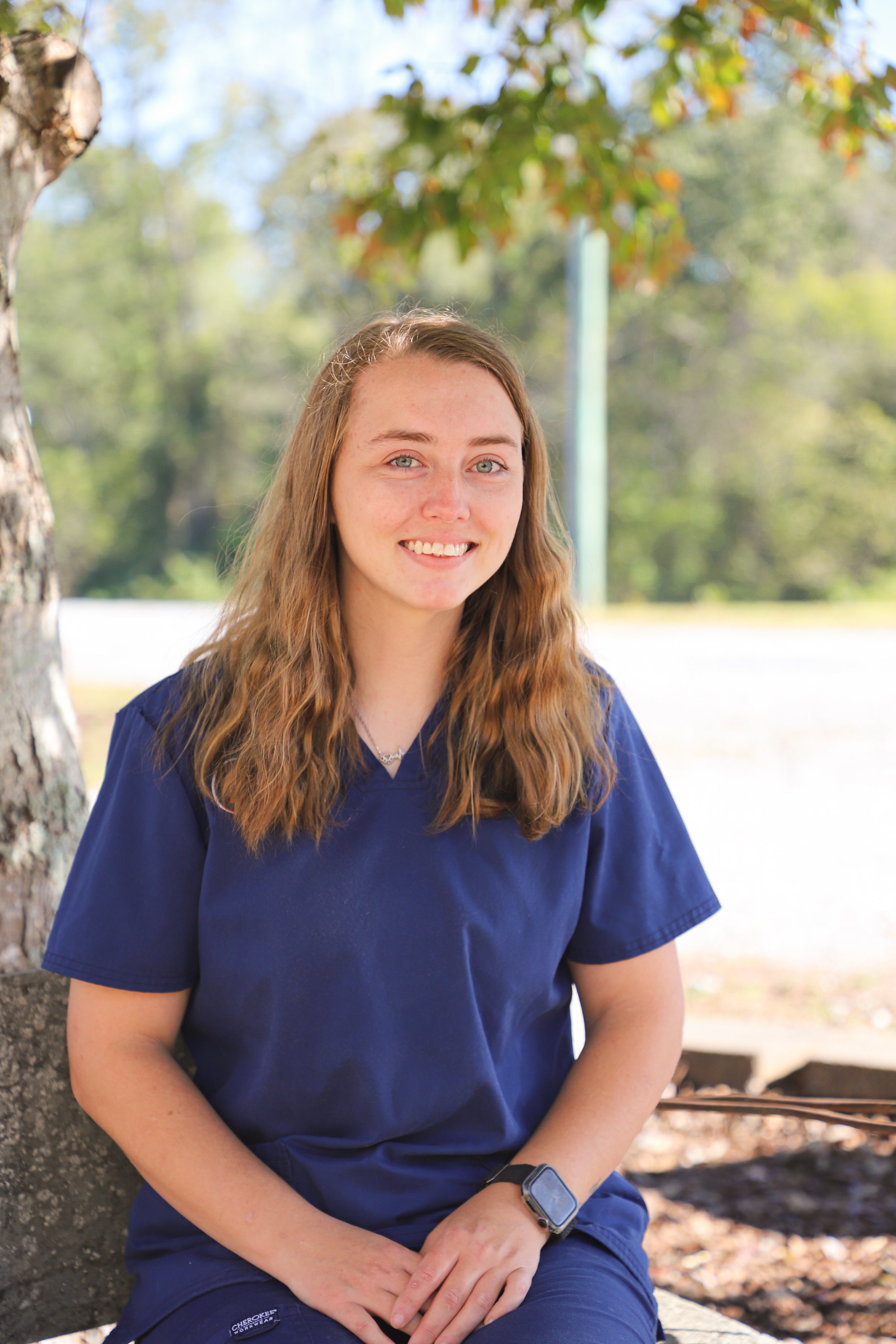 Young woman with long wavy light brown hair, wearing a navy blue uniform and a smartwatch, sitting outdoors on a bench under a tree.