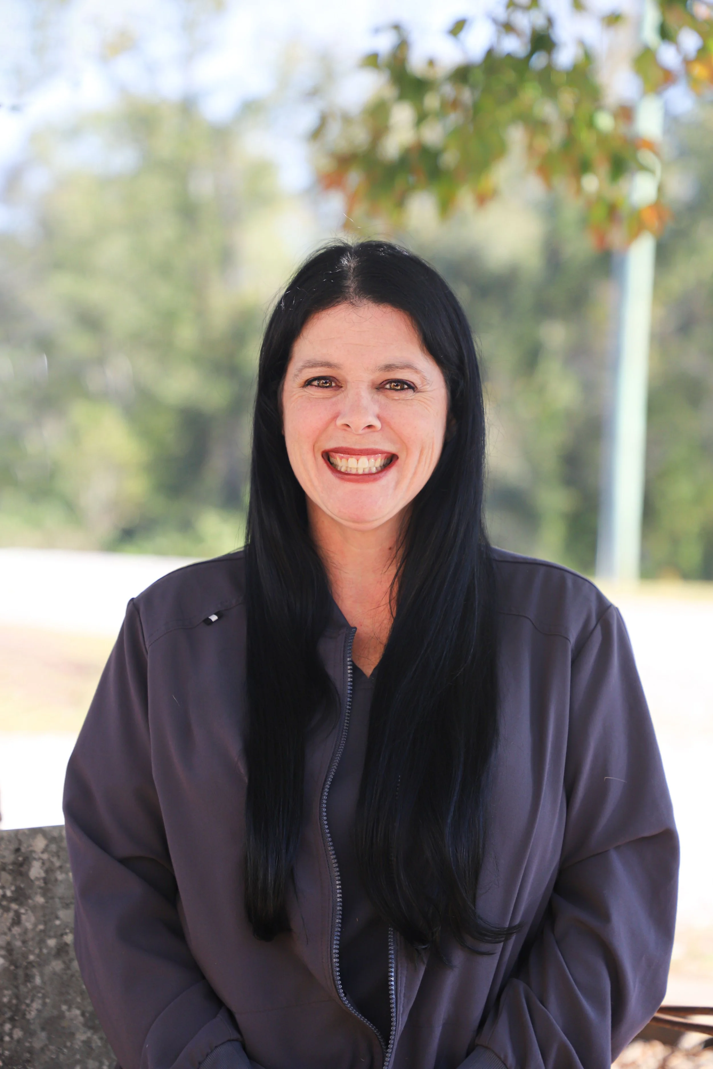 A woman with long black hair smiling outdoors with leaves and trees in the background.