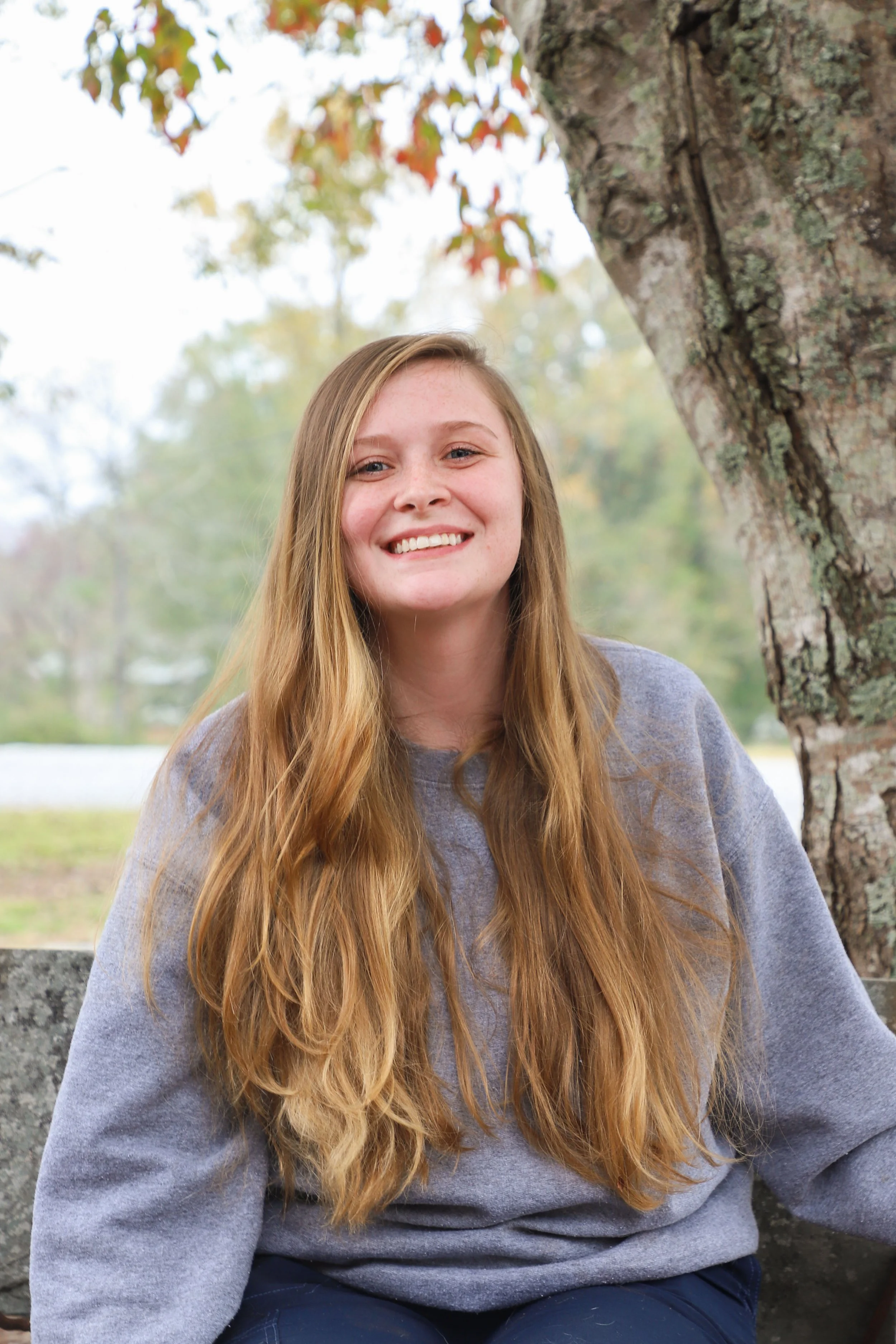 A young woman with long red hair, smiling, sitting outdoors near a tree with autumn leaves, wearing a gray sweatshirt.
