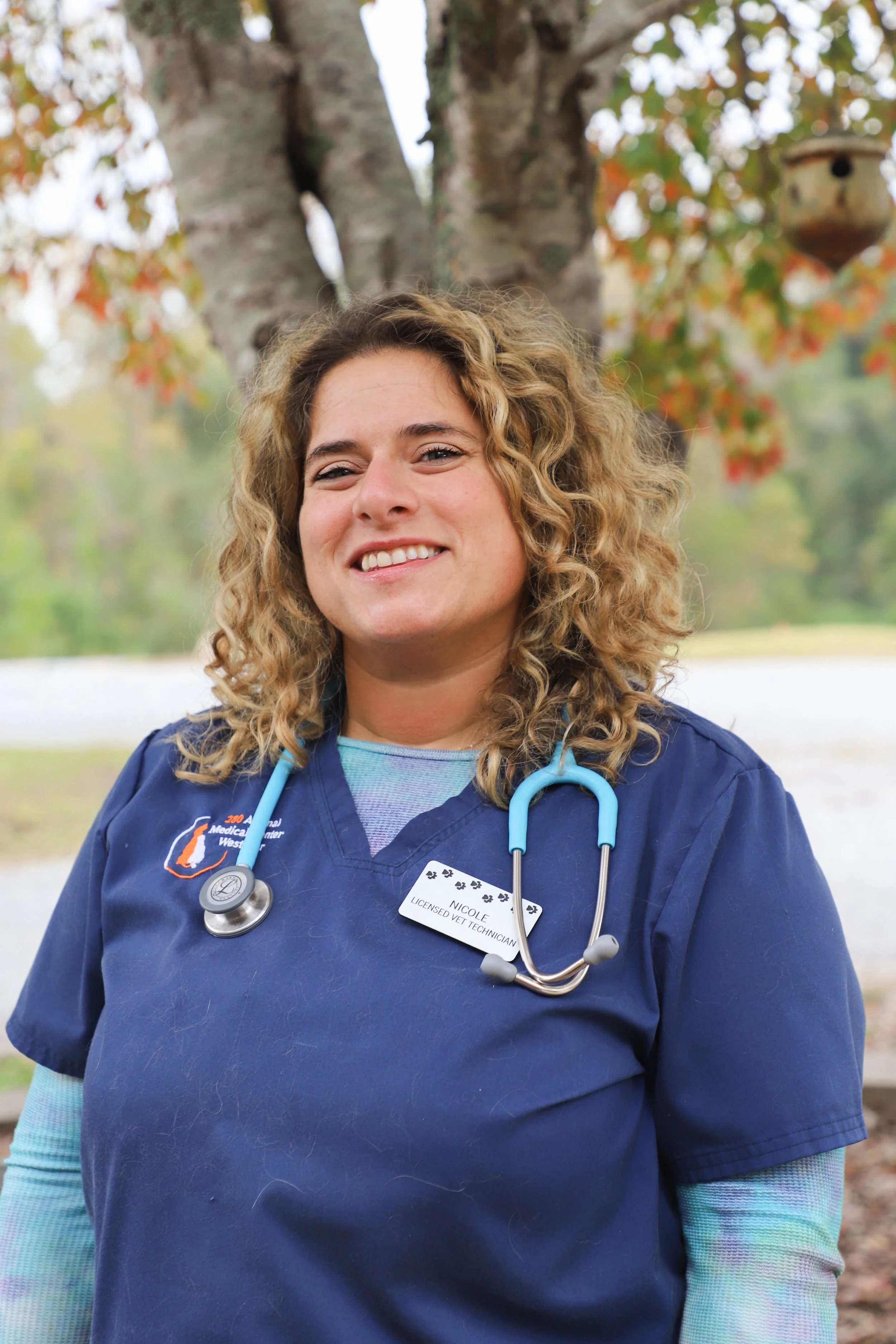 A woman in a blue nurse uniform with a stethoscope around her neck, smiling outdoors with a tree and fall foliage in the background.
