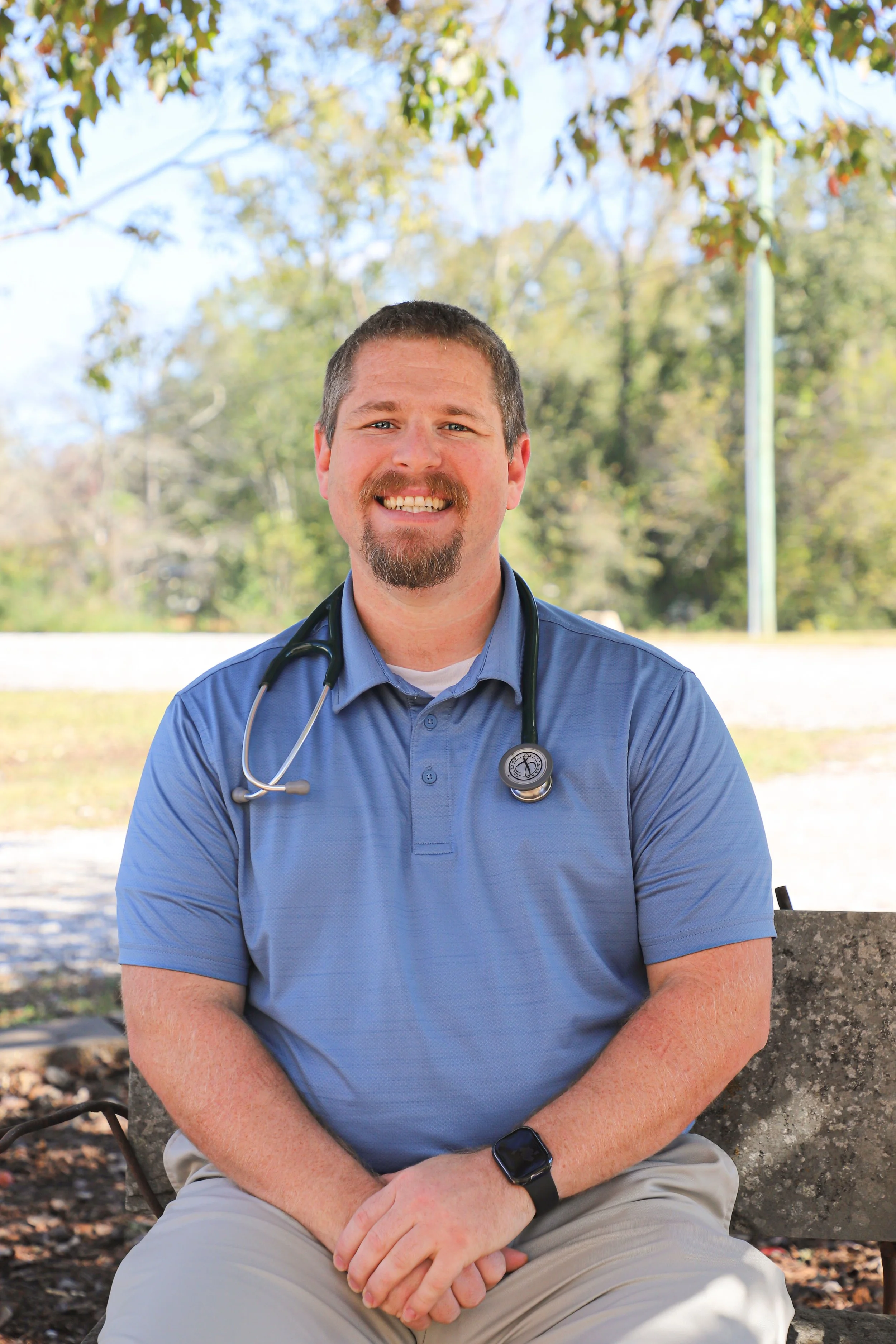 A man with short brown hair, a beard, and mustache smiling while sitting on a stone bench outdoors. He is wearing a blue polo shirt, a stethoscope around his neck, and a smartwatch on his left wrist. The background shows trees with green and some autumn-colored leaves, and a sunny sky.