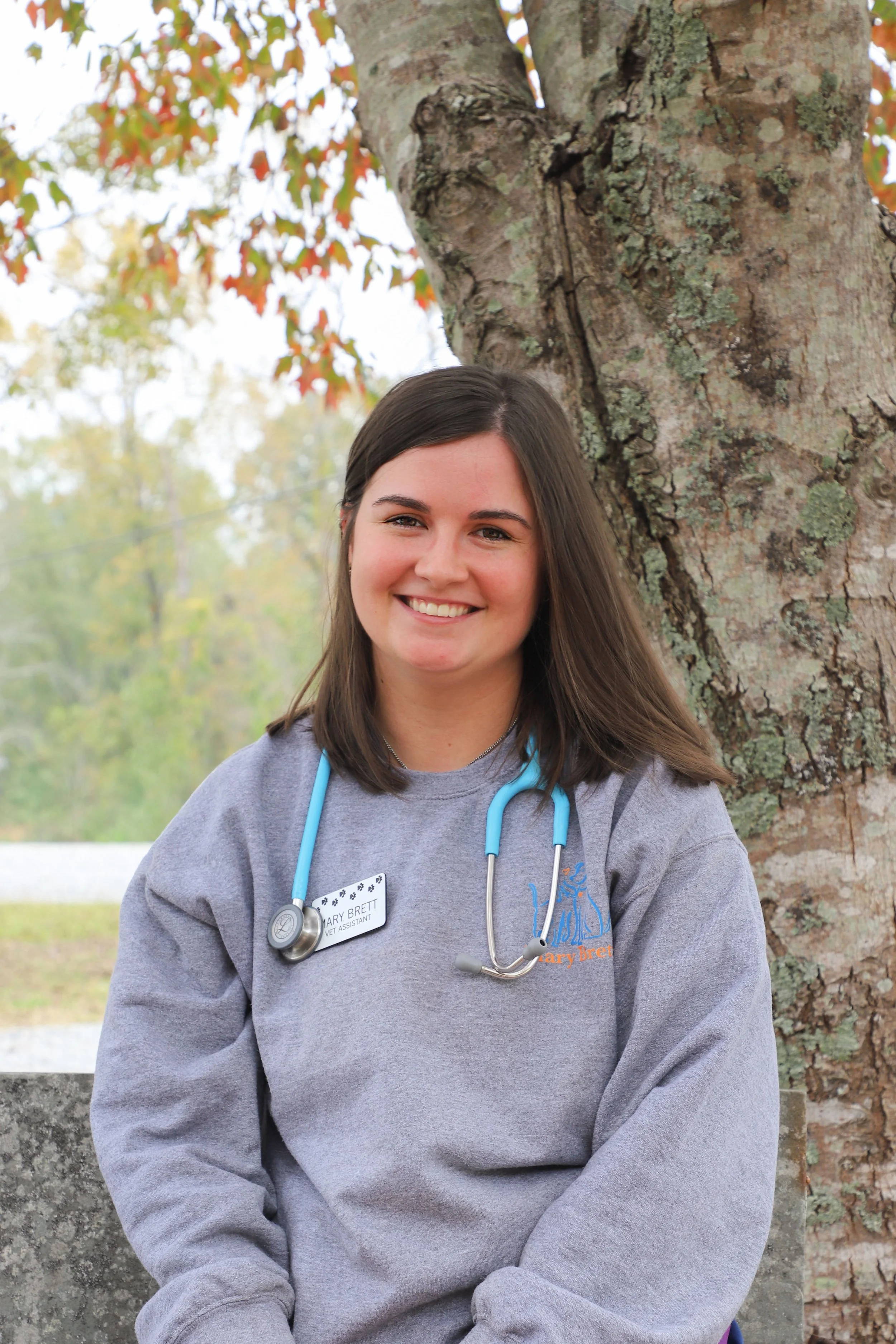 A smiling young woman with dark hair, wearing a gray sweatshirt and a stethoscope, standing outdoors next to a tree.