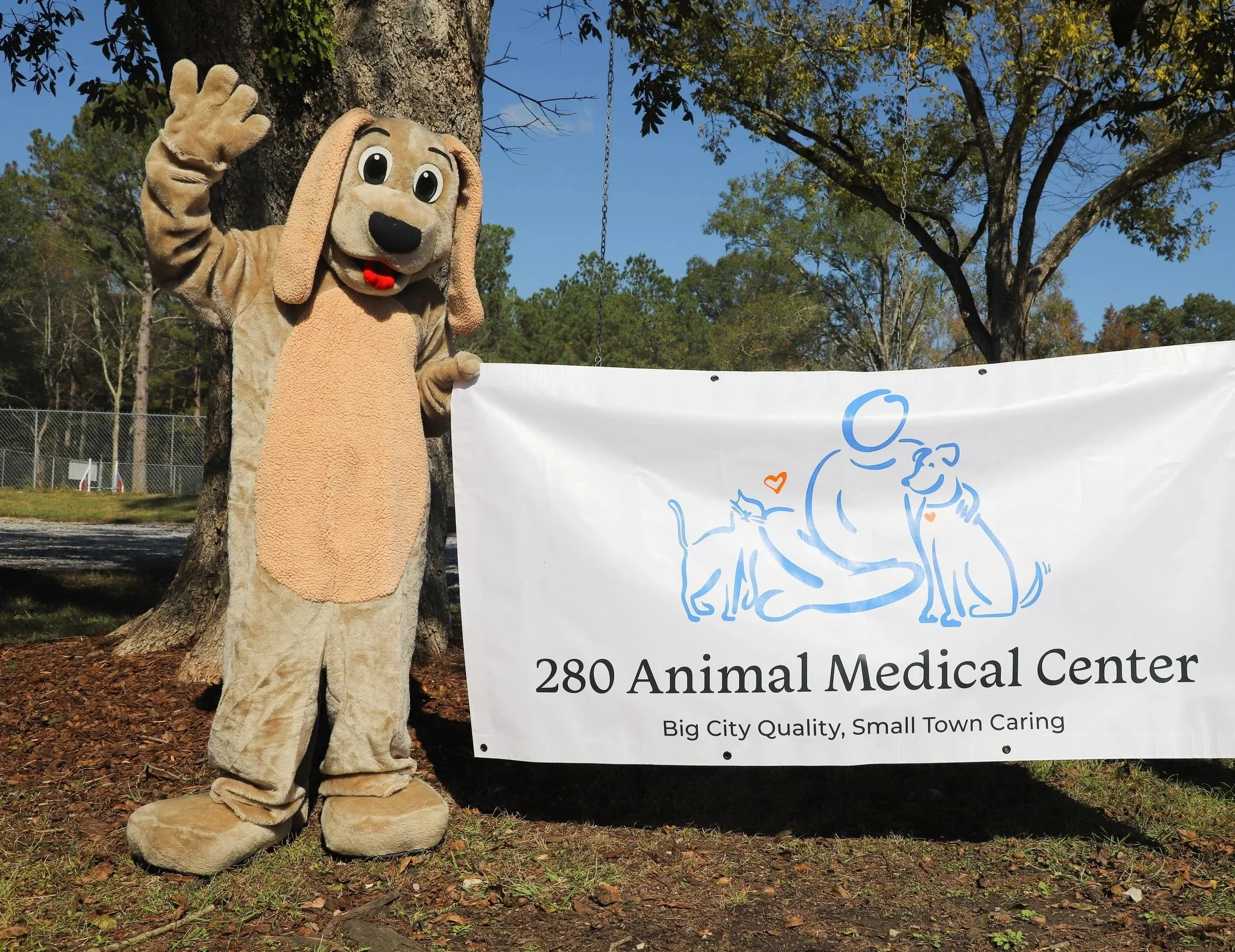 A person dressed in a dog mascot costume waving next to a banner that reads "280 Animal Medical Center." The banner features an illustration of a person with two dogs, and the tagline "Big City Quality, Small Town Caring." The scene is outdoors with trees and a fence in the background.