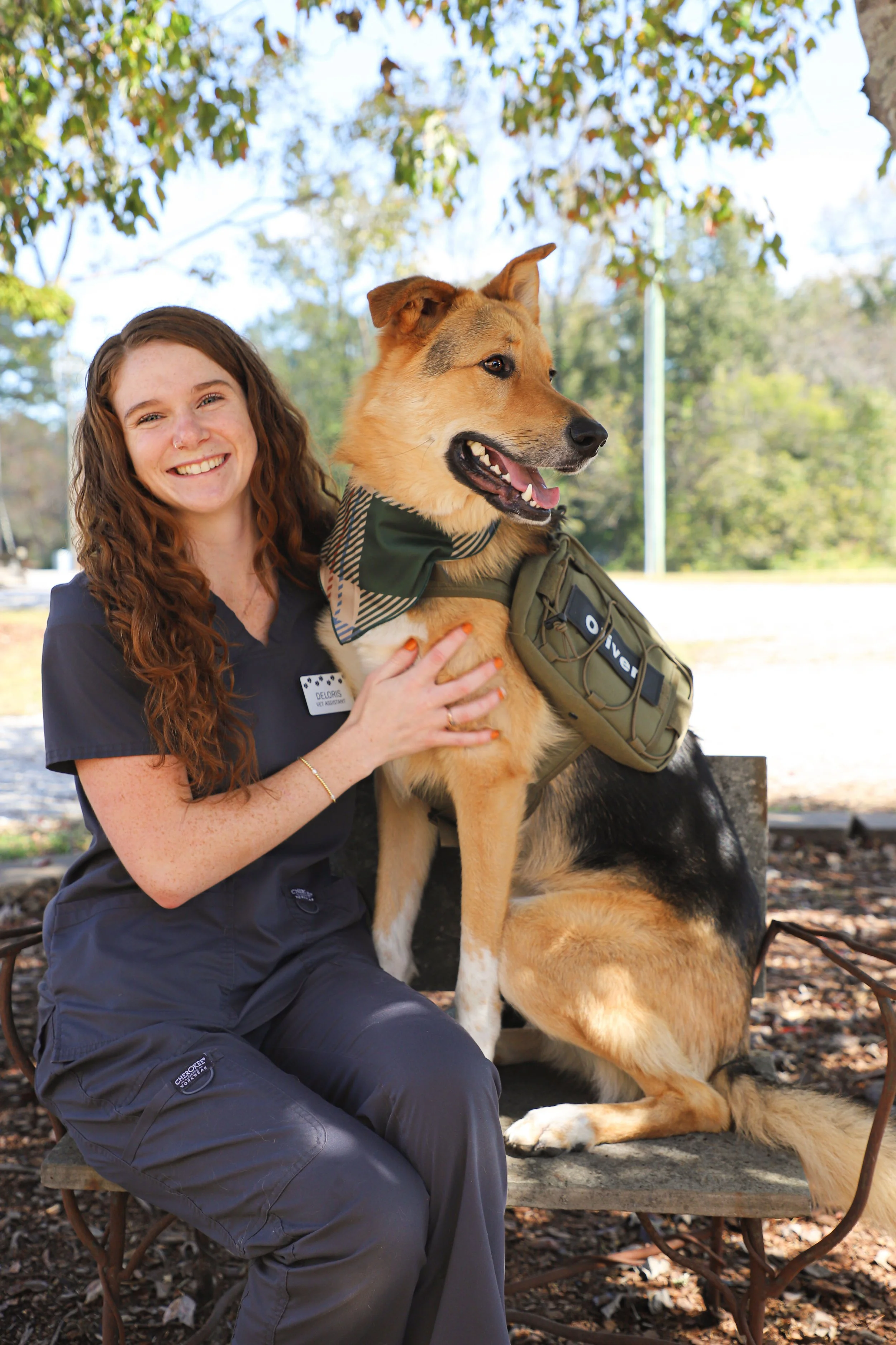 A woman with long, curly red hair wearing a dark gray shirt and matching pants, sitting on a park bench under a tree with green leaves, smiling while holding a large service dog with golden fur. The dog is wearing a tactical vest and bandana, sitting beside her outdoors on a bright, clear day.