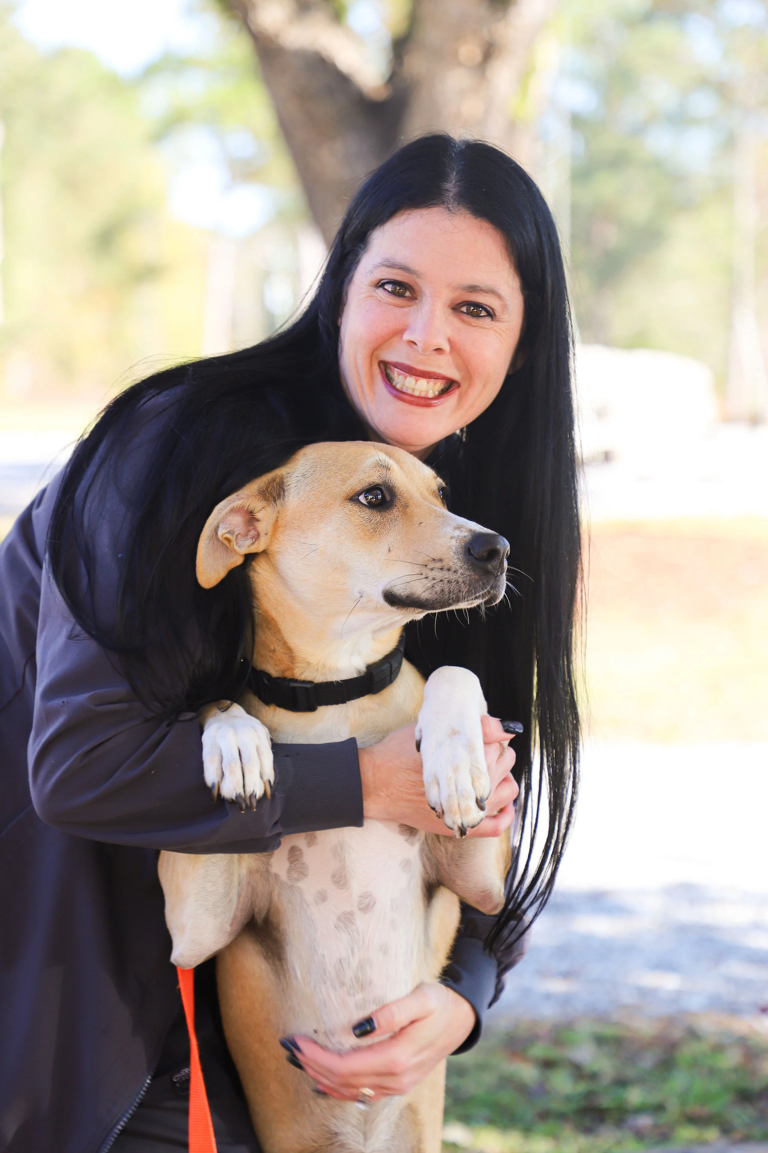A woman with long black hair smiling while holding a tan and white dog outdoors.