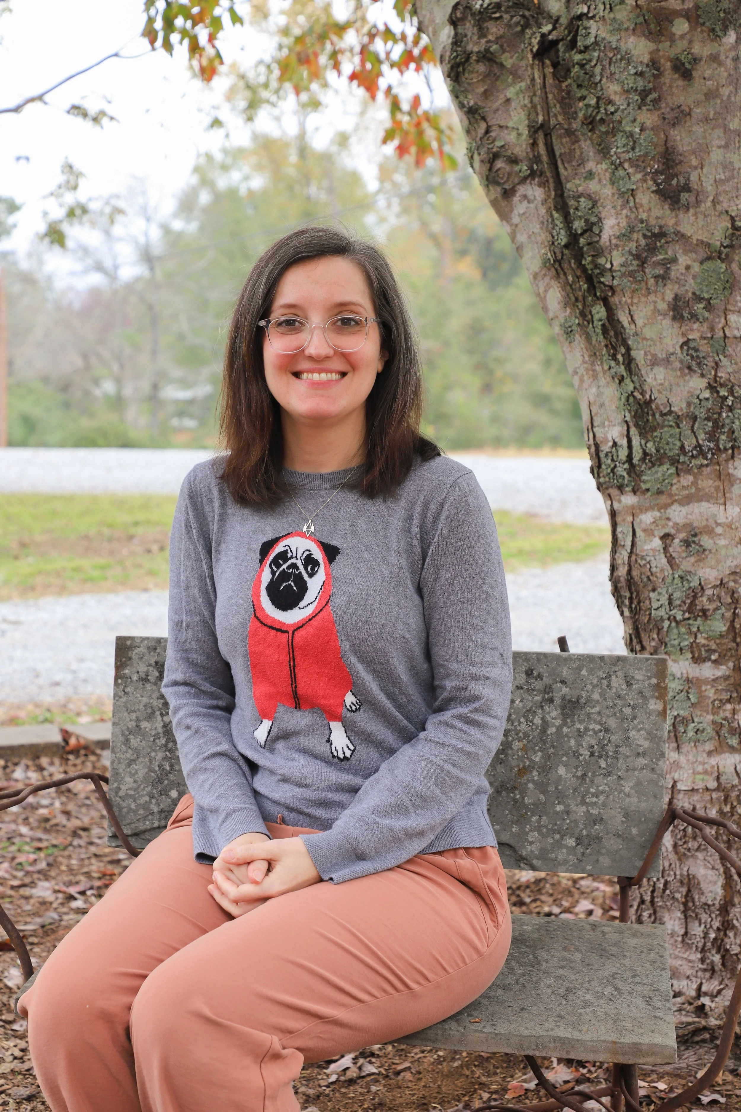A woman sitting on a park bench outdoors with a large tree next to her, wearing glasses, a gray sweater with a pug dog on it, and peach-colored pants, smiling at the camera.