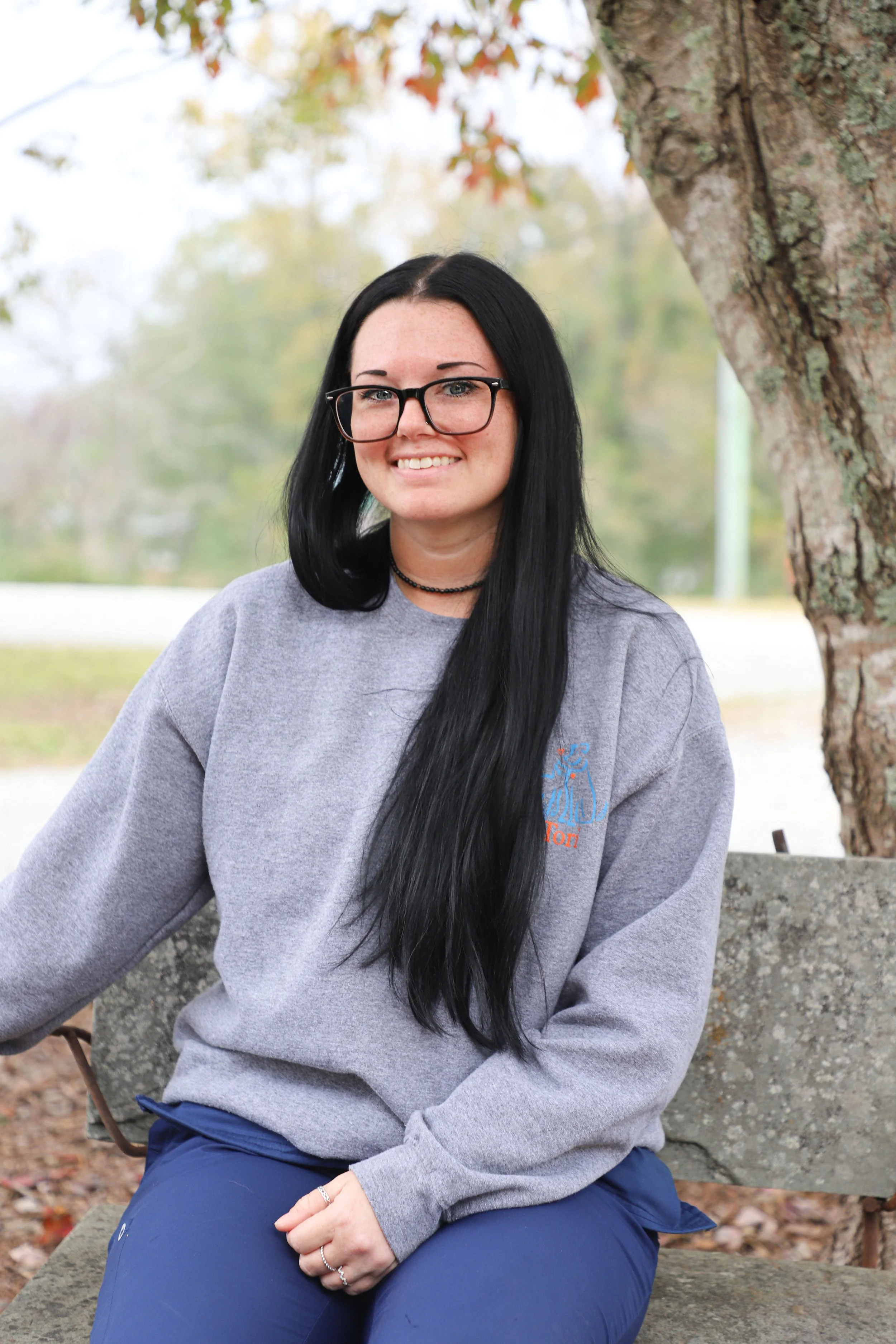 A woman with long black hair, glasses, and a black choker necklace sitting on a park bench outdoors, smiling, with a tree and autumn foliage in the background.