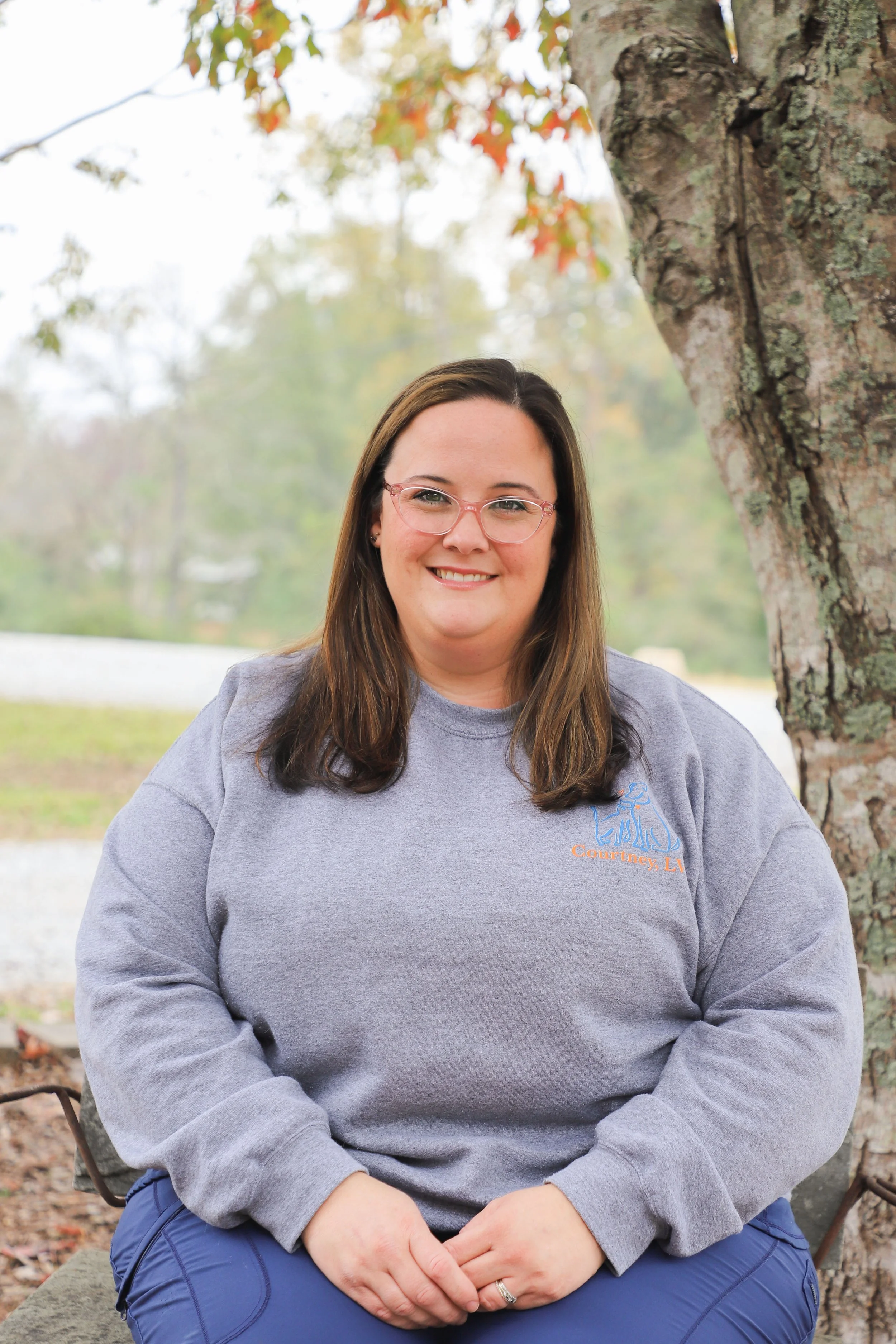 A woman with glasses, wearing a gray sweatshirt with orange and blue writing, seated outdoors next to a tree, smiling at the camera.