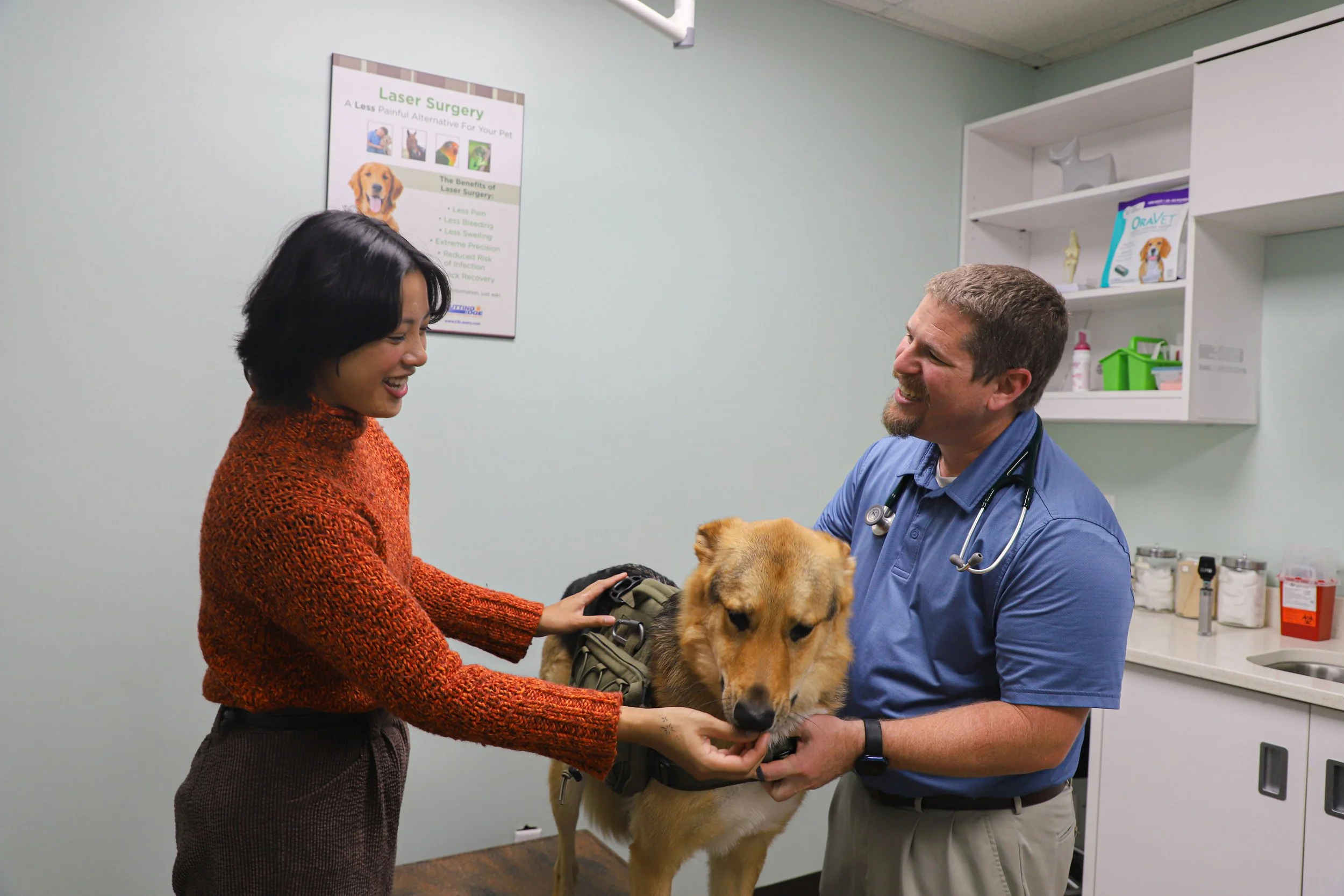 A woman and a veterinarian examining a golden retriever in a veterinary clinic. The woman is smiling and wearing an orange sweater, while the vet in blue scrubs has a stethoscope around his neck.