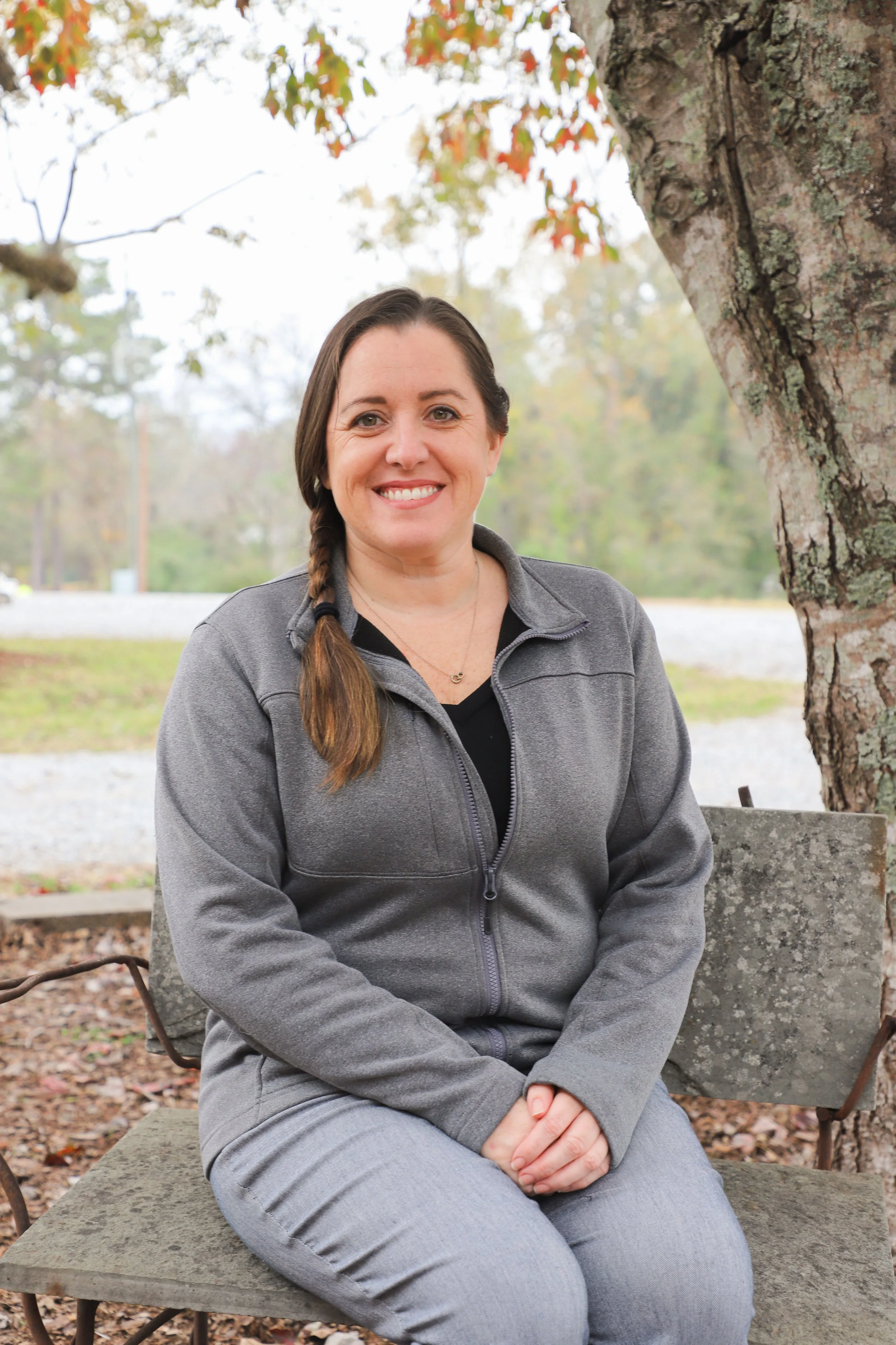 A woman with a braid, wearing a gray zip-up jacket and gray pants, sitting on a stone park bench outdoors under a tree with autumn foliage, smiling at the camera.