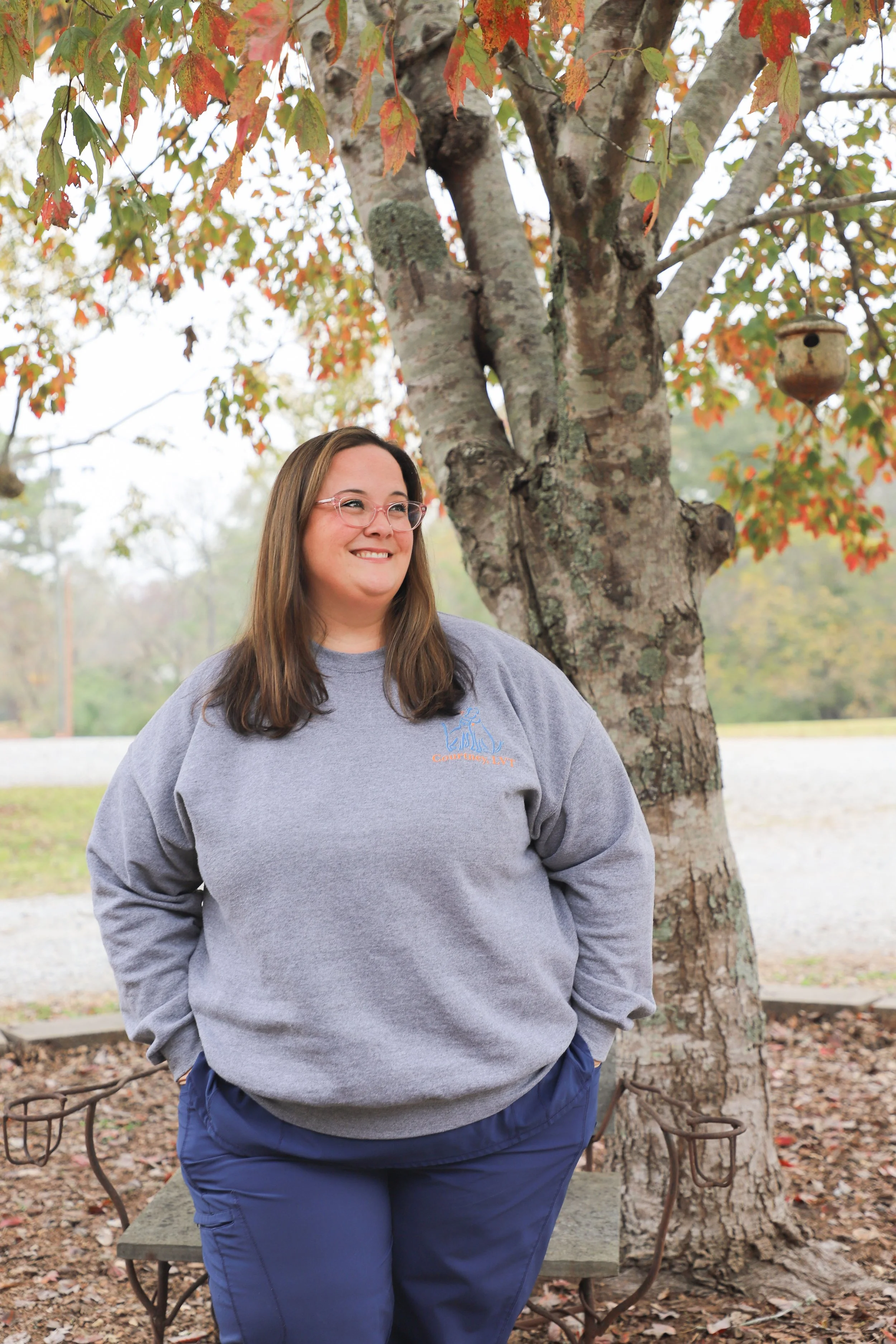 A woman with glasses and long brown hair standing outside in front of a tree with orange and green leaves, smiling and wearing a gray sweatshirt and blue pants.